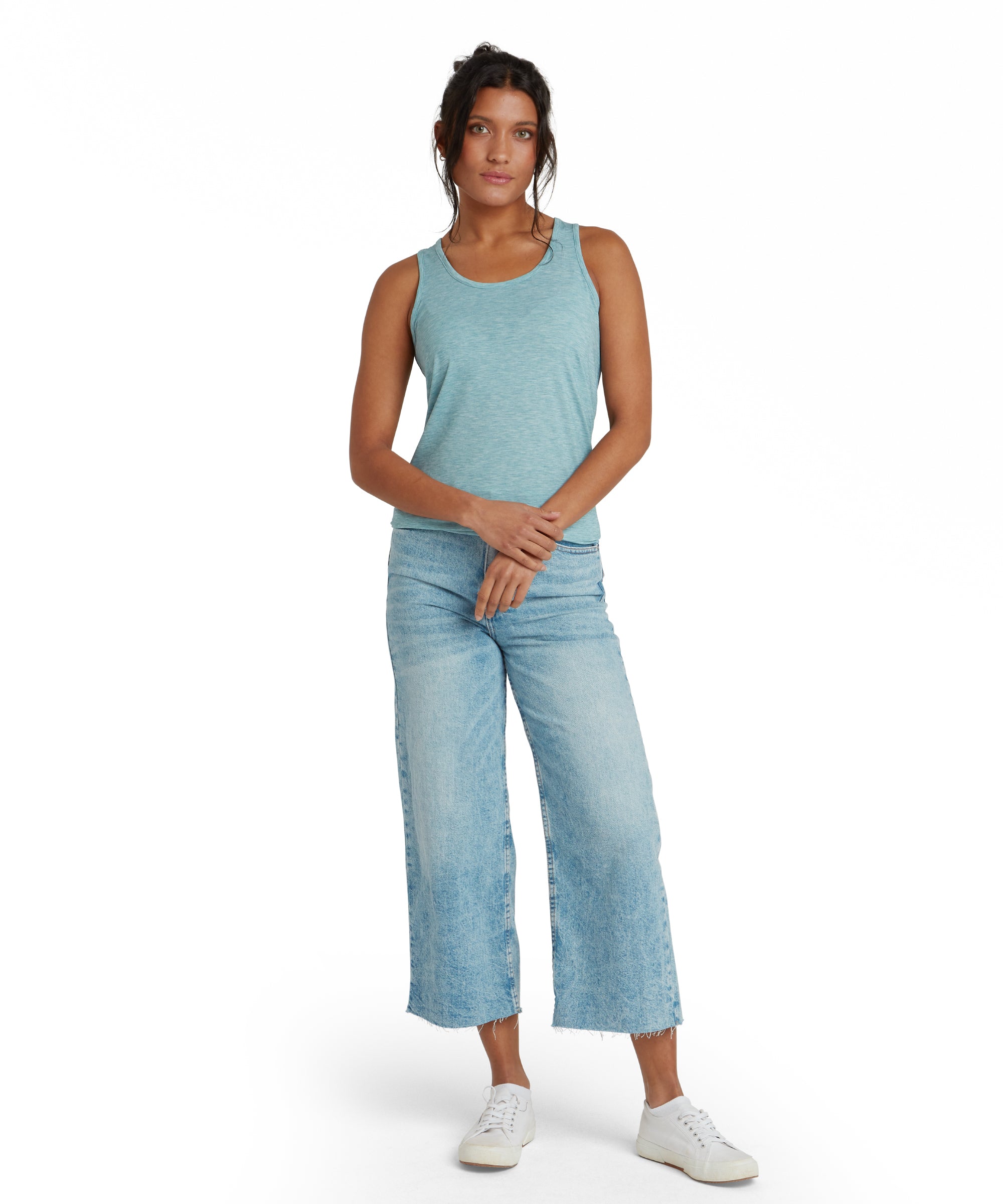 The woman is wearing a Sherpa Adventure Gear Asha Tank in Blue with a scooped neckline. The top has a lightweight, flowy fit and extends slightly below the hips. She has her hair tied back in a loose ponytail, and her expression is calm and confident.