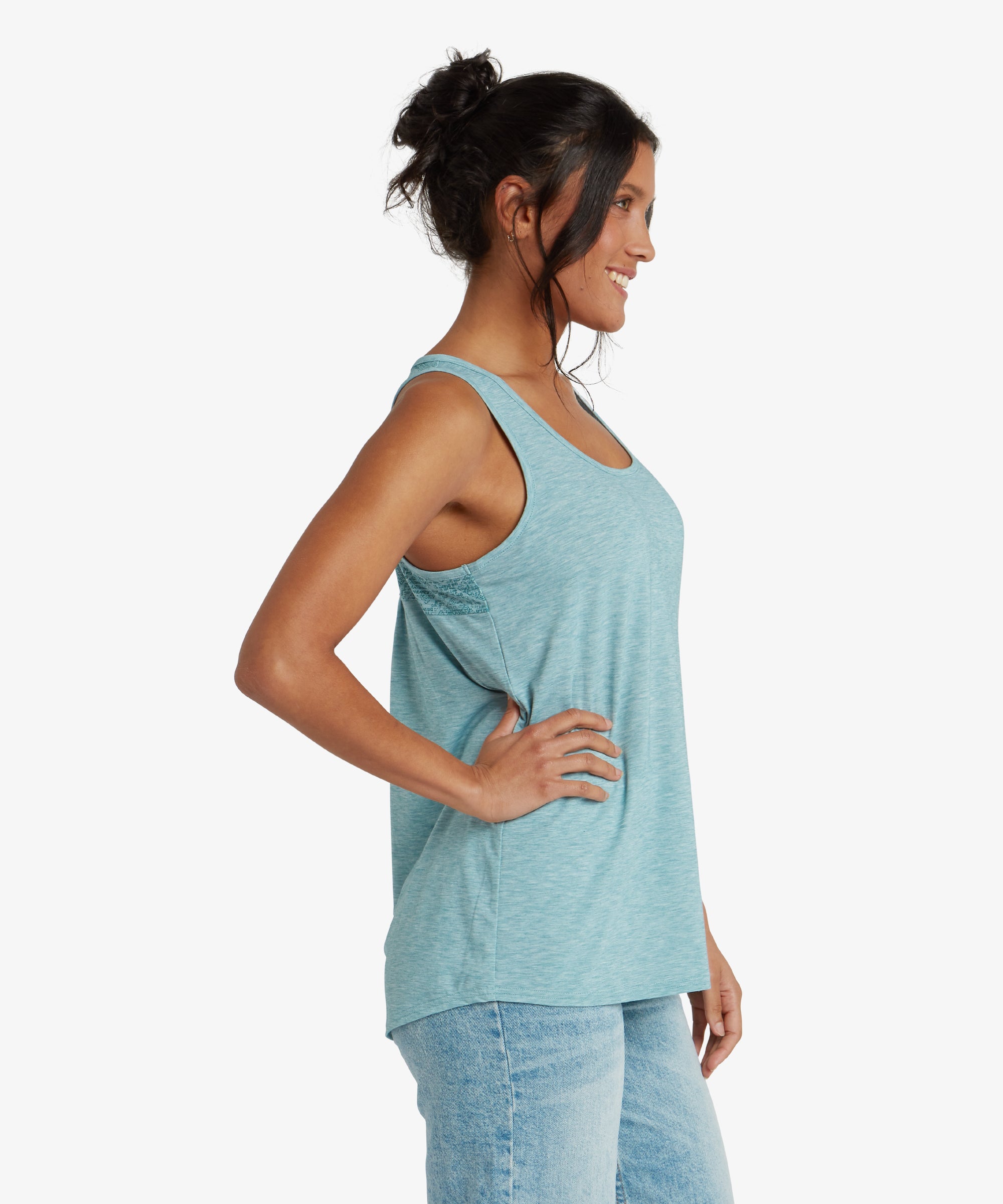 The woman is captured from the side, showcasing the relaxed drape of the Sherpa Adventure Gear Asha Tank in Blue. The armholes are cut moderately low, revealing the top's breathable design. The fabric has a subtle heathered pattern, emphasising its casual and comfortable style.