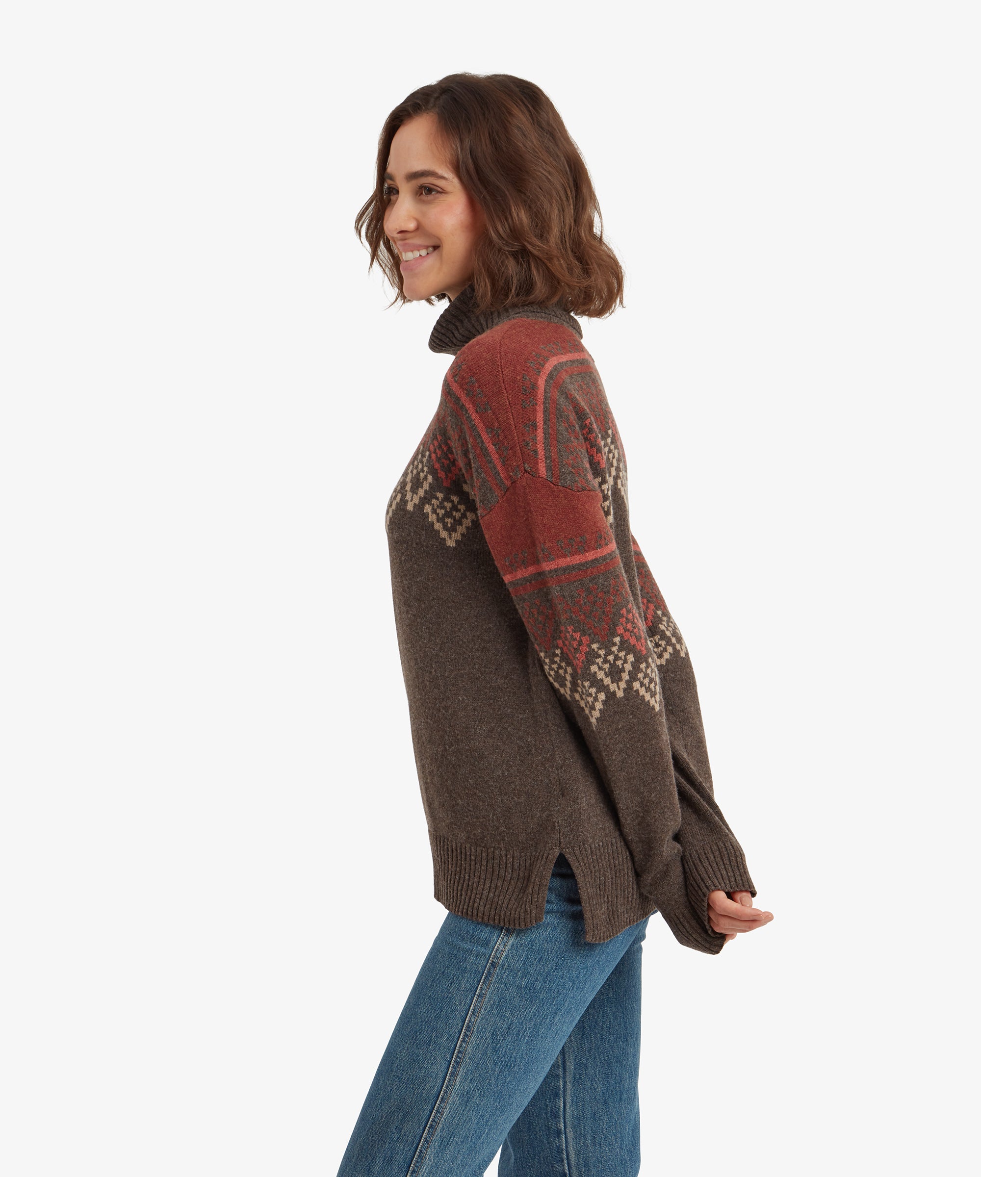 A side profile view of a woman wearing a Sherpa Adventure Gear Azha Roll Neck Sweater in Brown with red and beige patterns on the sleeves and shoulders. She is smiling, with the loose hem of the sweater and side slits visible, paired with light blue jeans.