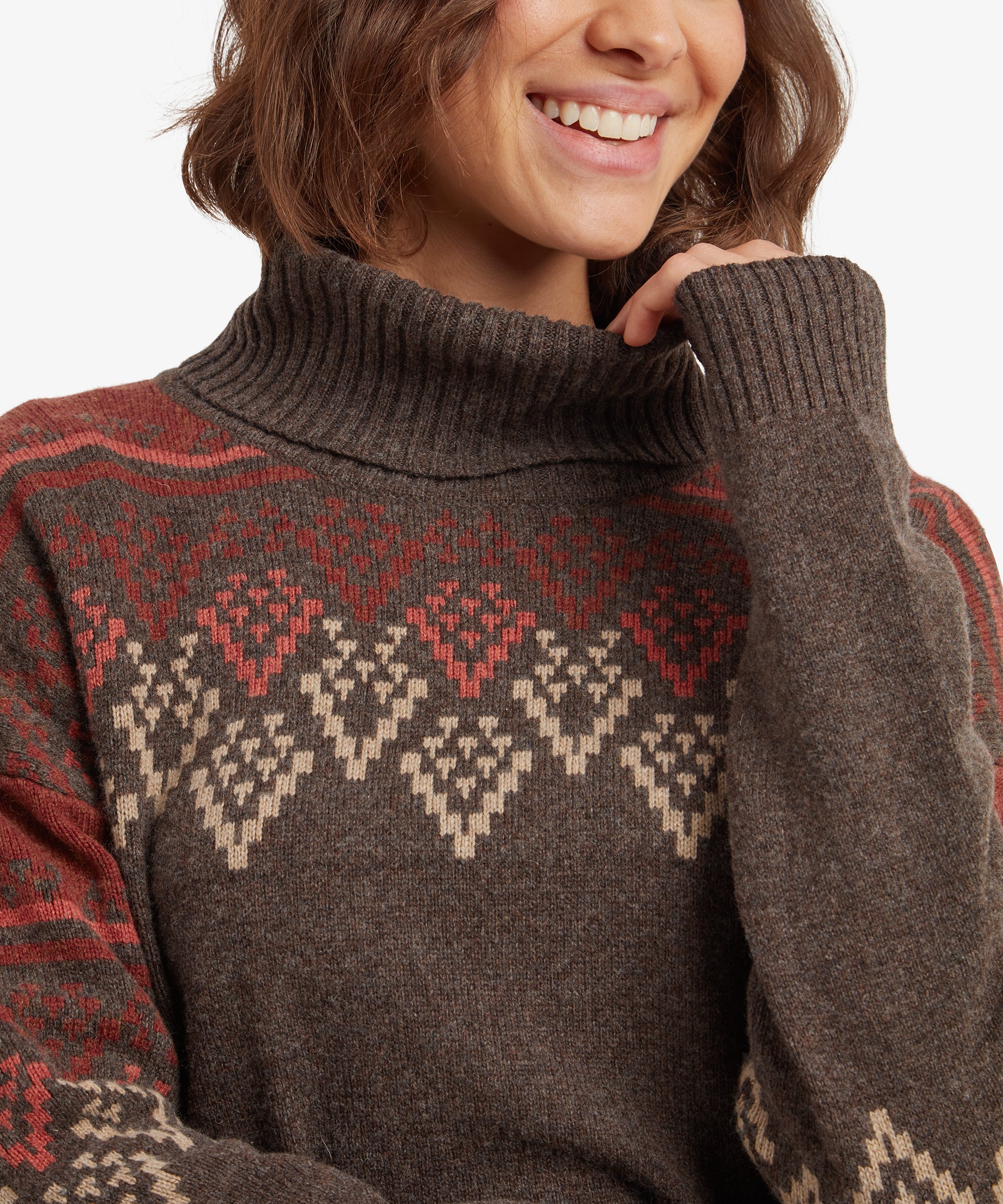 A close-up of the Sherpa Adventure Gear Azha Roll Neck Sweater in Brown's neckline, highlighting the textured roll-neck design and the detailed diamond patterns in beige and red. The woman’s smile is visible in the frame.