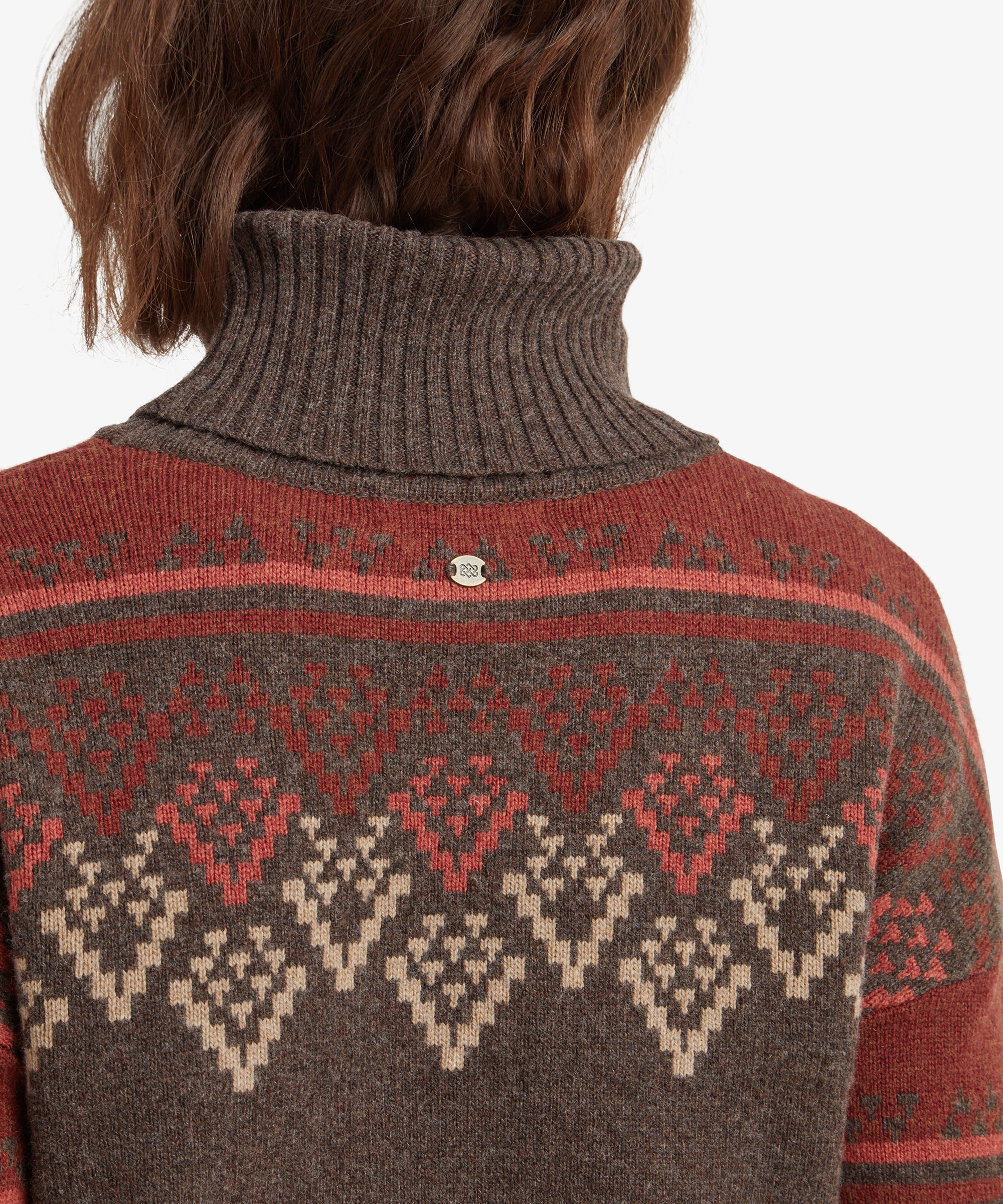 A close-up view of the back of the Sherpa Adventure Gear Azha Roll Neck Sweater in Brown's roll neck, focusing on the textured knit pattern and small decorative metal tag on the upper back. The intricate red and beige diamond design is also prominent in the background.