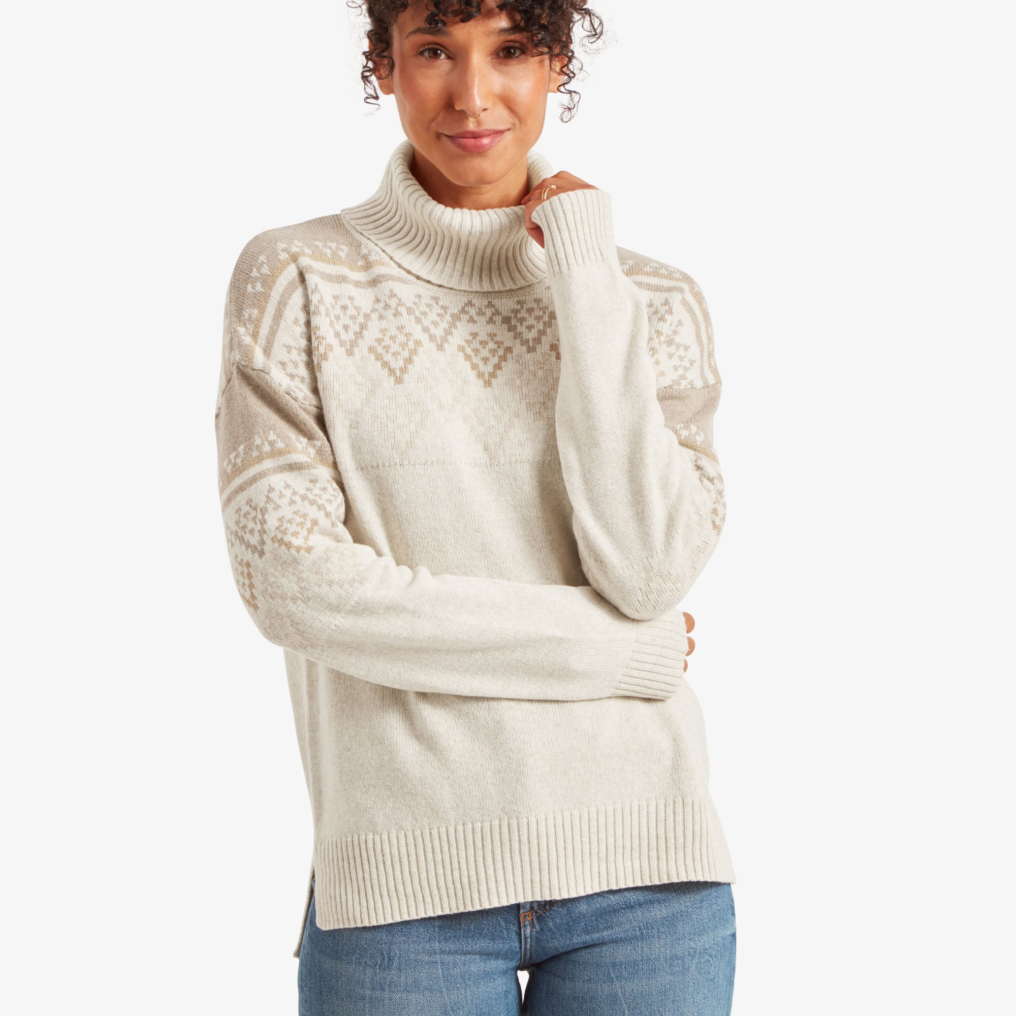 A woman is wearing a Sherpa Adventure Gear Azha Roll Neck Sweater in White with intricate fair isle-inspired diamond patterns in beige tones. She is gently holding the neck of the sweater with one hand, smiling softly. The sweater features ribbed cuffs and hem, creating a cozy look. She pairs it with light blue denim.