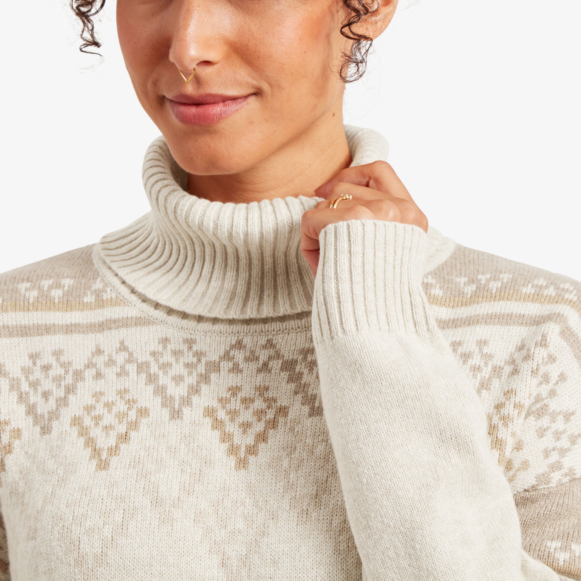 A detailed look at the Sherpa Adventure Gear Azha Roll Neck Sweater in White's roll-neck design and intricate beige diamond patterns. The ribbed texture of the neck contrasts nicely with the smooth pattern, and the soft woolen material is apparent. The model's hands are lightly touching the neckline.