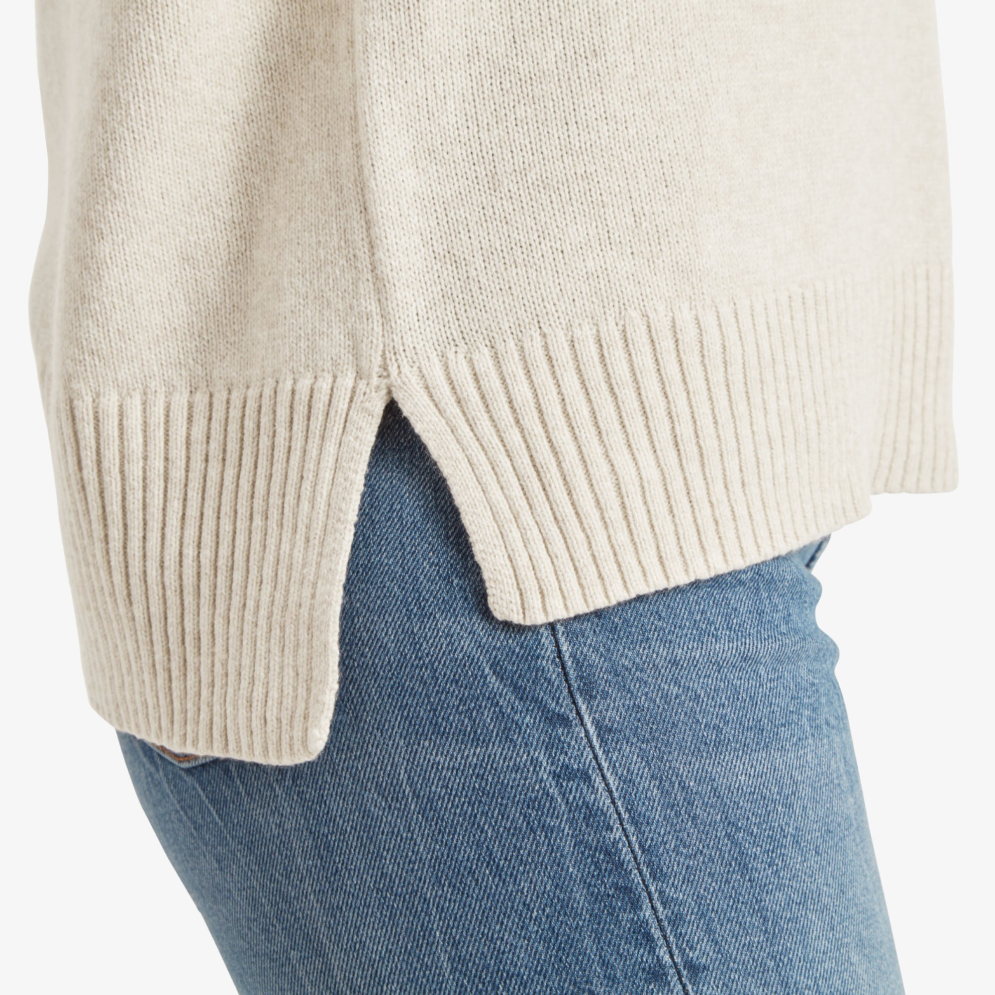 A close-up of the side hem of the Sherpa Adventure Gear Azha Roll Neck Sweater in White, featuring a small side slit for added comfort and mobility. The ribbed hem is thick and contrasts slightly with the smooth body of the sweater. The hem rests over light blue jeans, showing the pairing.