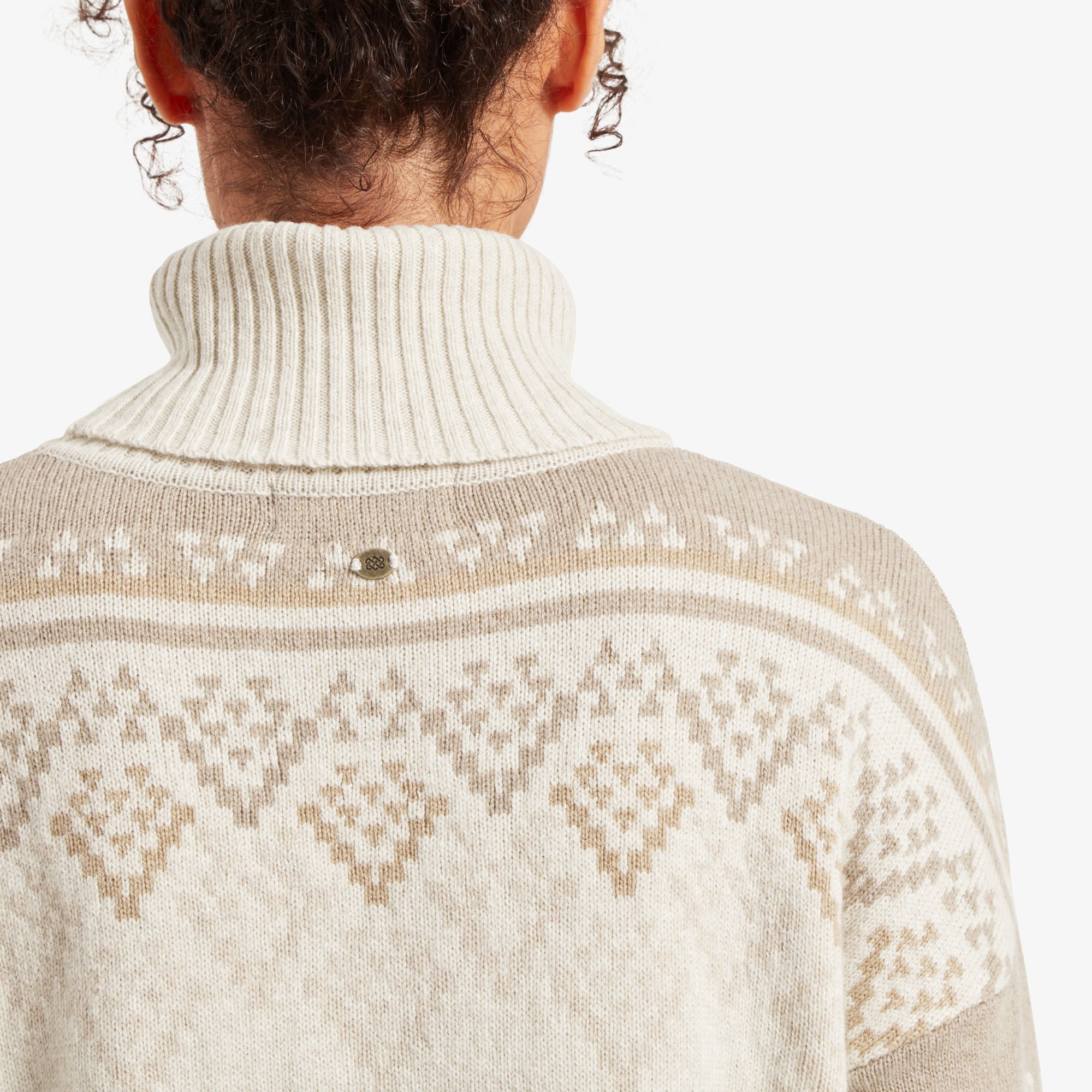 A close-up view of the back neckline of the Sherpa Adventure Gear Azha Roll Neck Sweater in White, showing a small, branded metallic emblem centered beneath the rolled neck. The intricate diamond pattern is prominently displayed, highlighting the craftsmanship of the knit design.