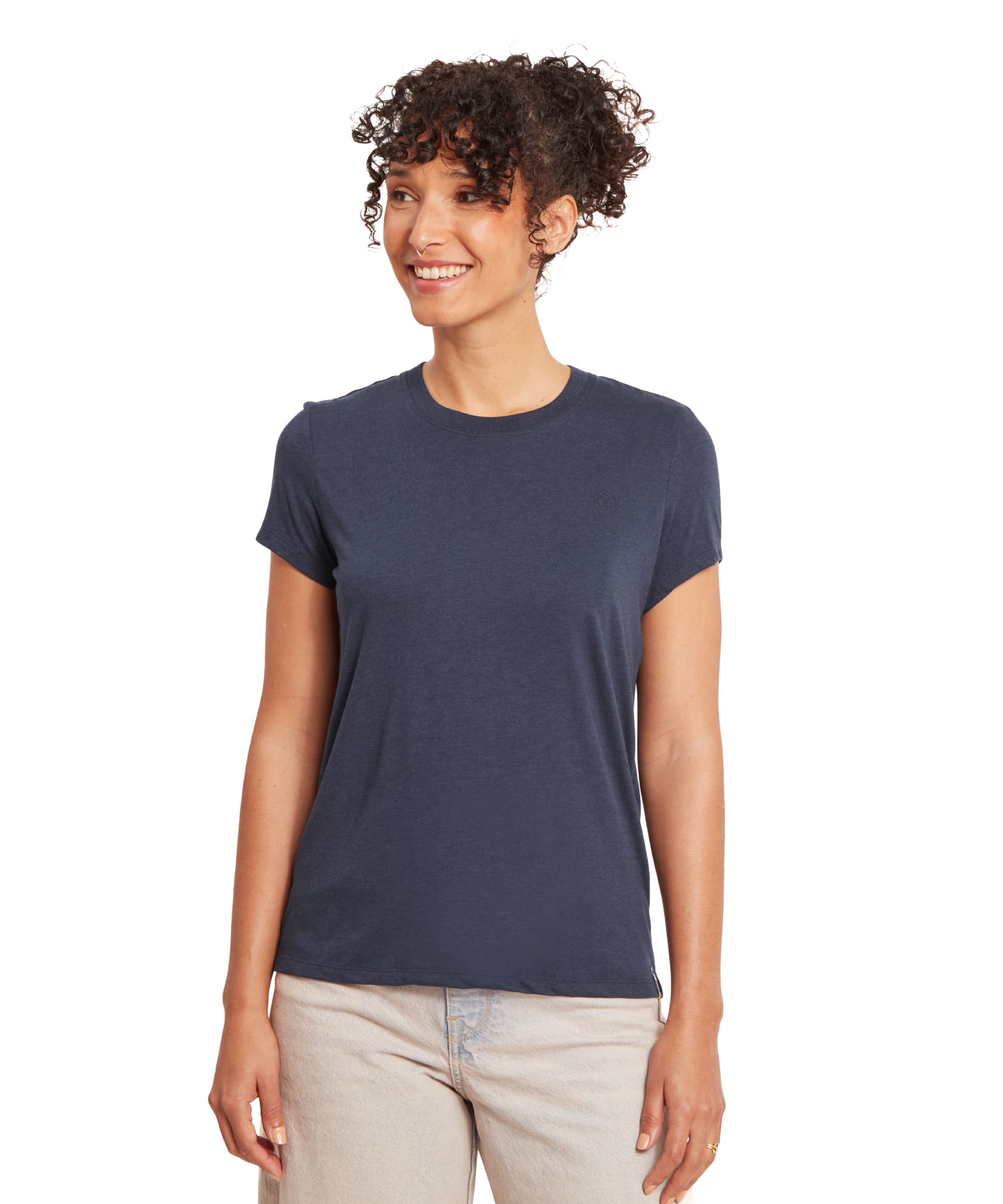 A woman wearing a Sherpa Adventure Gear Bali T-Shirt in Blue styled casually with beige pants. The t-shirt features a minimalist design with a small embroidered Sherpa logo in navy on the left side of the chest. The woman is smiling and standing upright, showcasing the relaxed fit of the shirt.