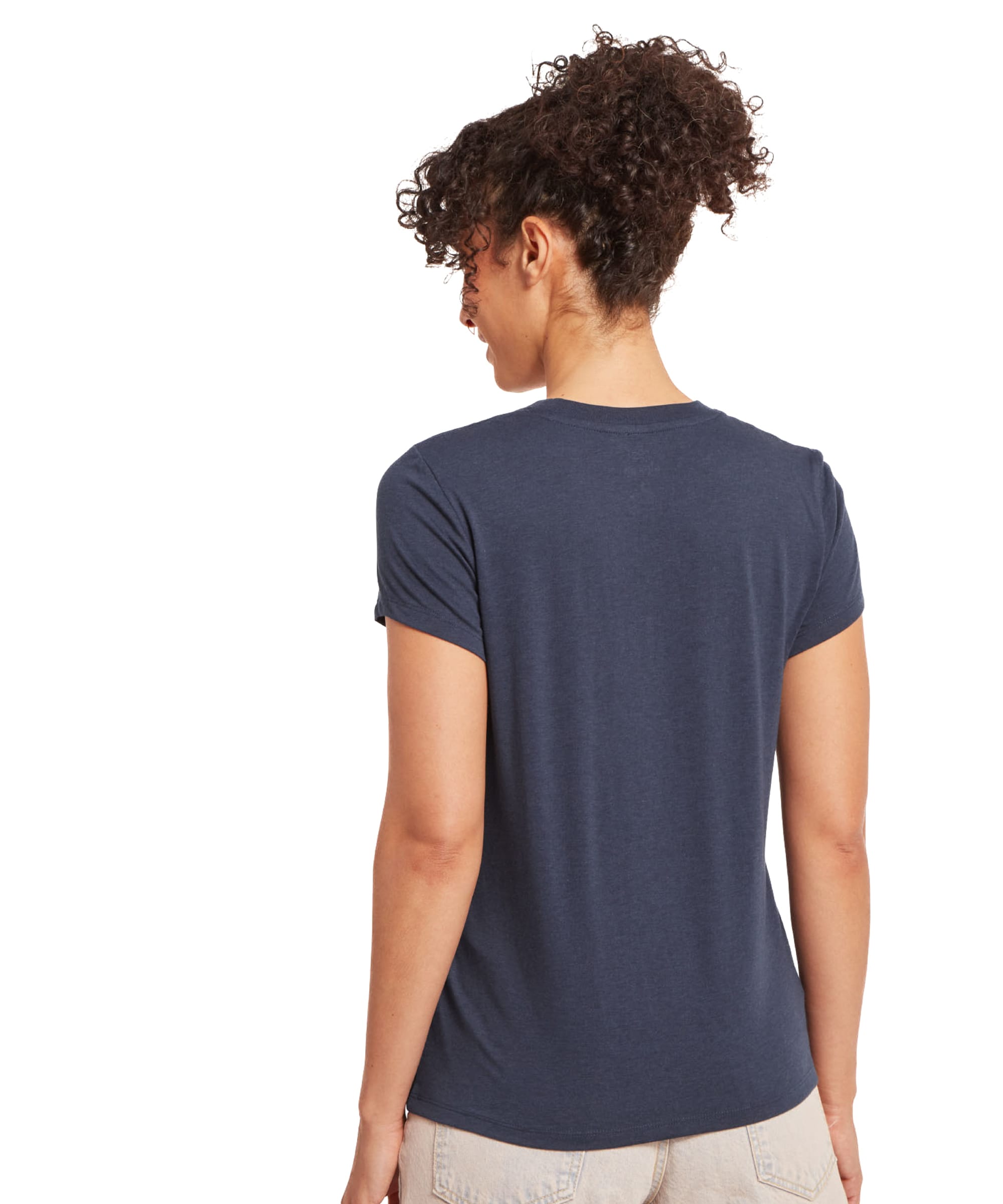 The same woman is turned around, displaying the back of the Sherpa Adventure Gear Bali T-Shirt in Blue. The fabric is smooth, with no visible patterns or graphics, and the hemline sits neatly over beige pants. Her curly hair is tied up, emphasising the simplicity of the t-shirt's design.