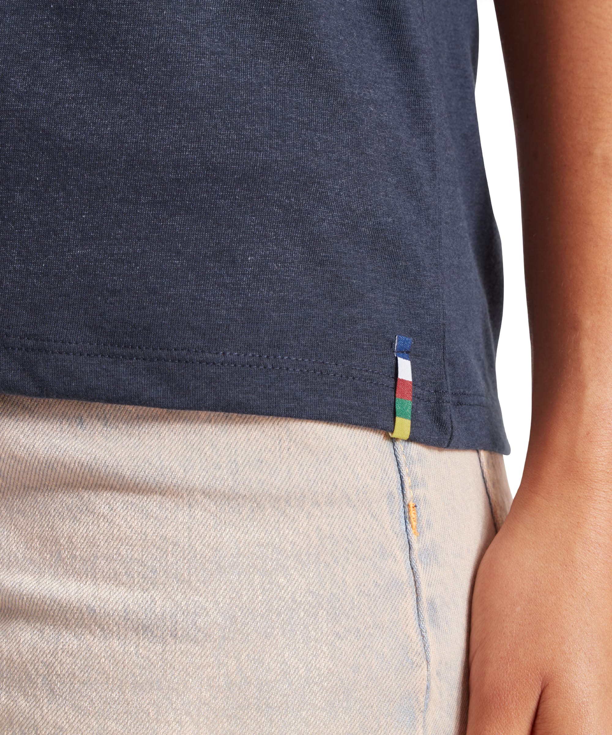 A close-up view of the bottom hem of the Sherpa Adventure Gear Bali T-Shirt in Blue, where a small multicolored fabric tag in blue, red, green, and yellow is stitched into the seam. The image also captures part of the beige pants, providing a clear view of the fine stitching and relaxed fit.