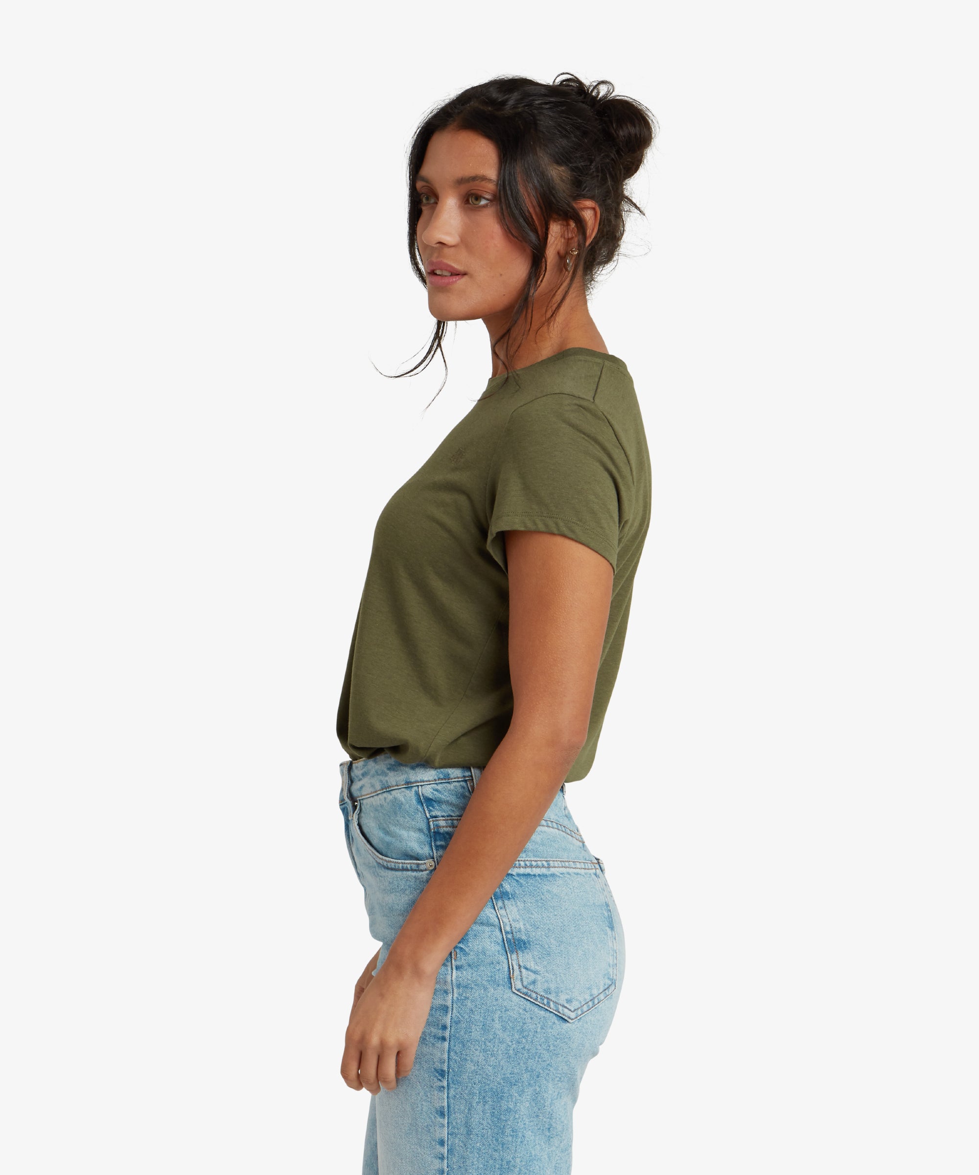 A rear view of the model showcasing the back of the Sherpa Adventure Gear Bali T-Shirt in Green. The T-shirt has a slightly curved hemline and a smooth fit. Her wavy ponytail cascades down her back, and she gently tilts her head to the side.