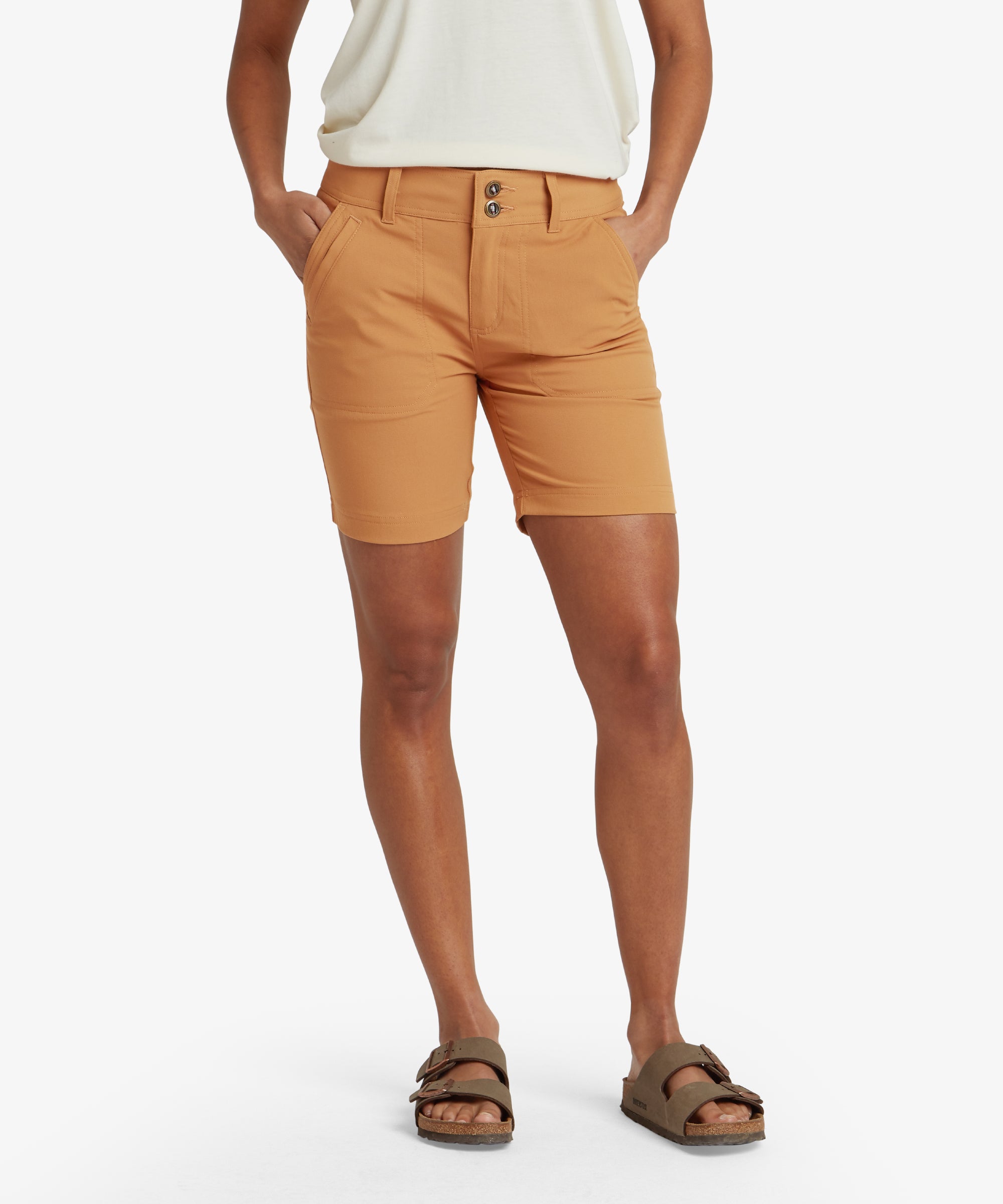 A front view of a woman wearing Sherpa Adventure Gear Bara Shorts in Light Brown. The shorts feature a double-button closure at the waistband and side pockets. The model pairs the shorts with white trainers and a tucked-in white t-shirt, showcasing a clean and casual look.