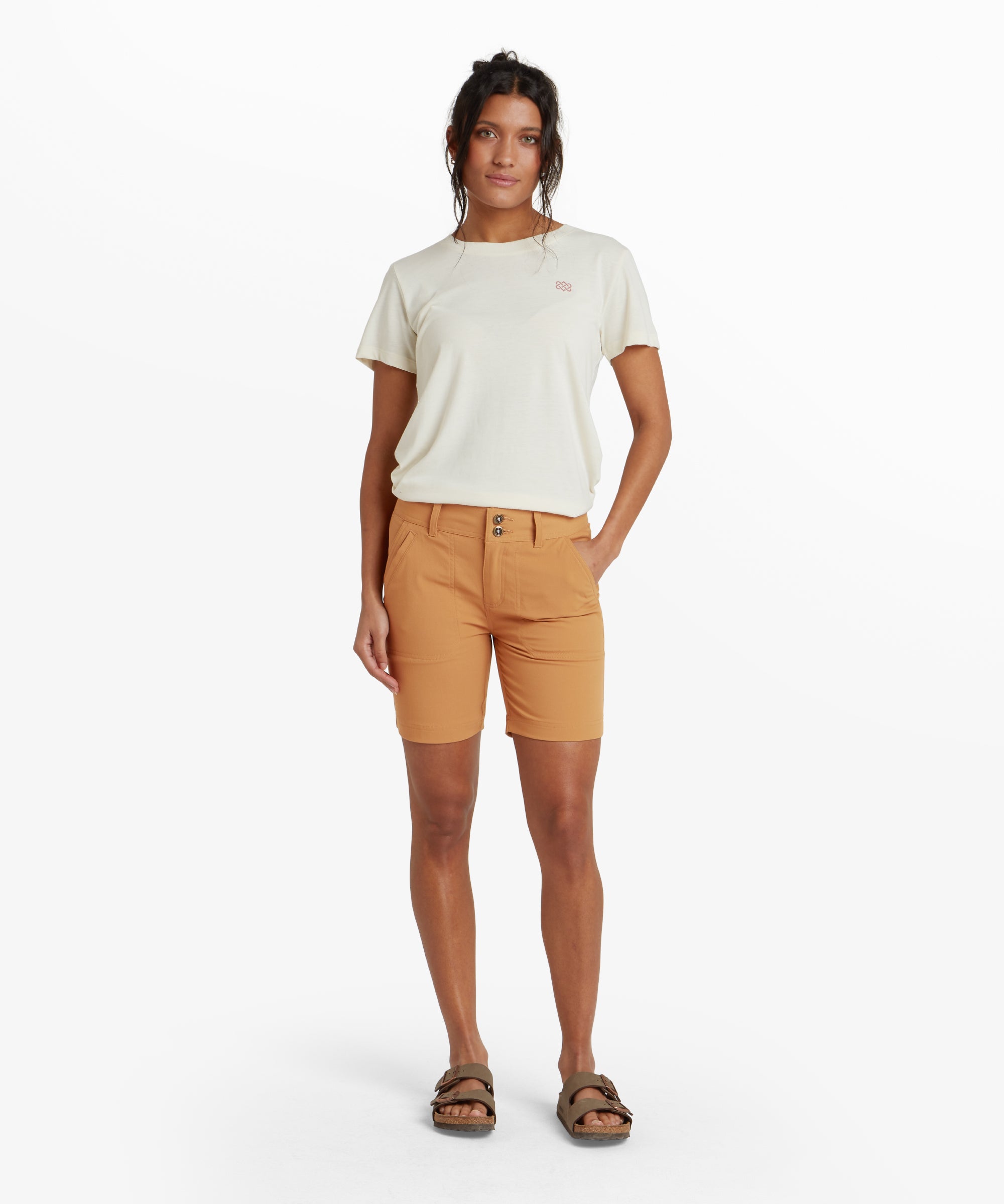 A full-body image of the model standing confidently while wearing Sherpa Adventure Gear Bara Shorts in Light Brown with a double-button closure. She complements the shorts with a graphic t-shirt and white trainers. The neutral background emphasises the shorts’ tailored fit and clean design.