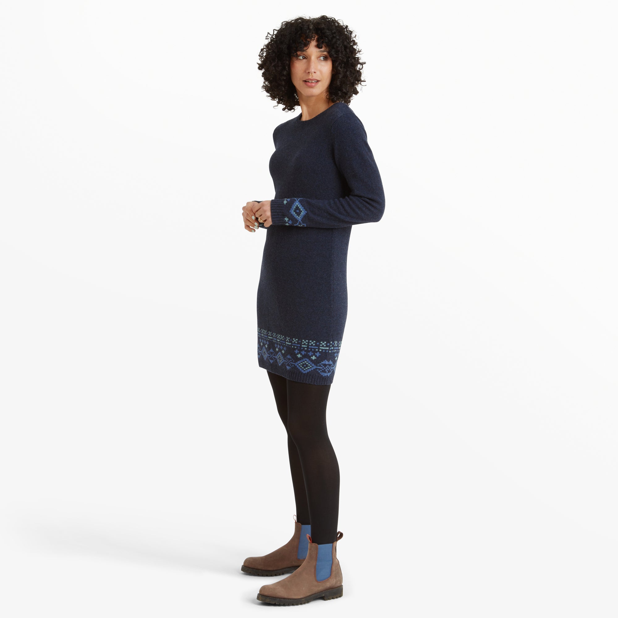 A side view of the same woman wearing the Sherpa Adventure Gear Bharati Eco Dress in Blue, showcasing the garment's silhouette and length. The geometric patterned details on the cuffs and hemline are clearly visible. Her outfit is paired with black tights and the same brown and blue boots, emphasising a casual yet elegant look.