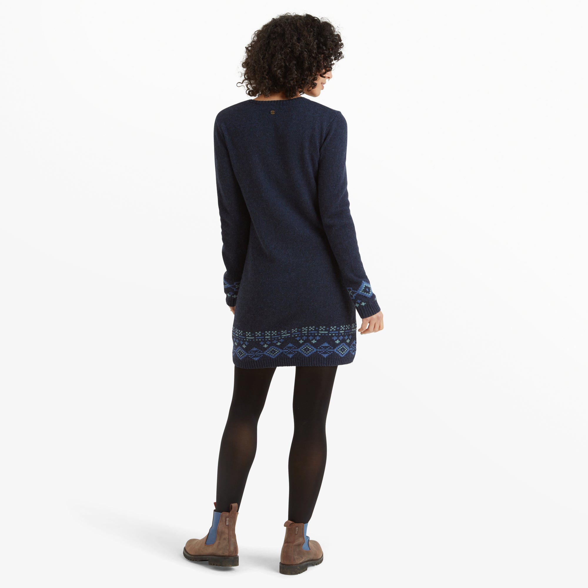 A rear view of the Sherpa Adventure Gear Bharati Eco Dress in Blue, highlighting the back's simplicity and the continuation of the geometric patterned details on the hemline and cuffs. The woman’s curly hair frames her face as she turns slightly to the side, giving a full perspective of the garment's design.