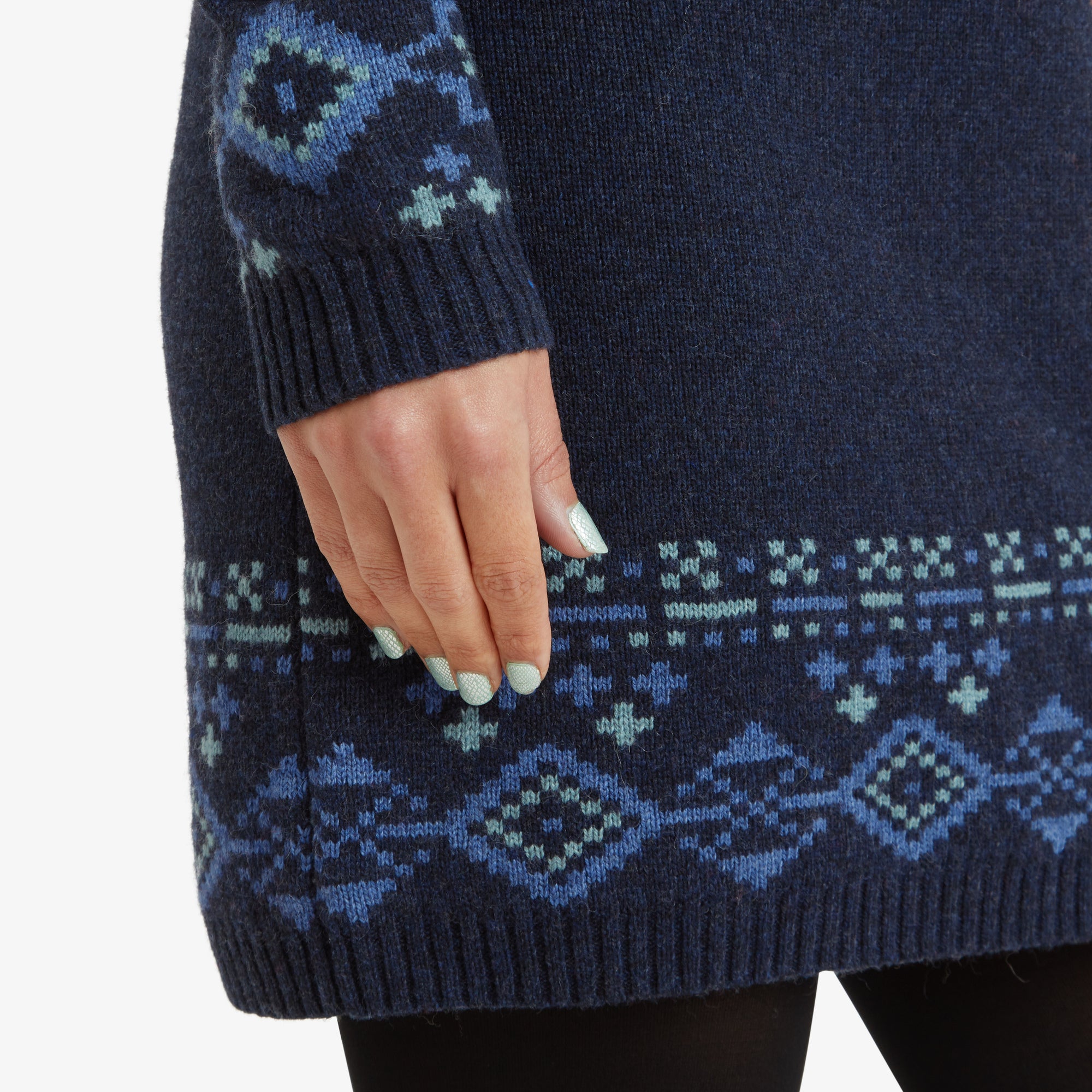 A close-up shot of the hemline and sleeve cuff detailing of the Sherpa Adventure Gear Bharati Eco Dress in Blue. The intricate blue and teal geometric patterns are prominently displayed, along with the soft texture of the knit fabric. The model's hand rests gently on the dress, with her light green painted nails complementing the outfit's color scheme.