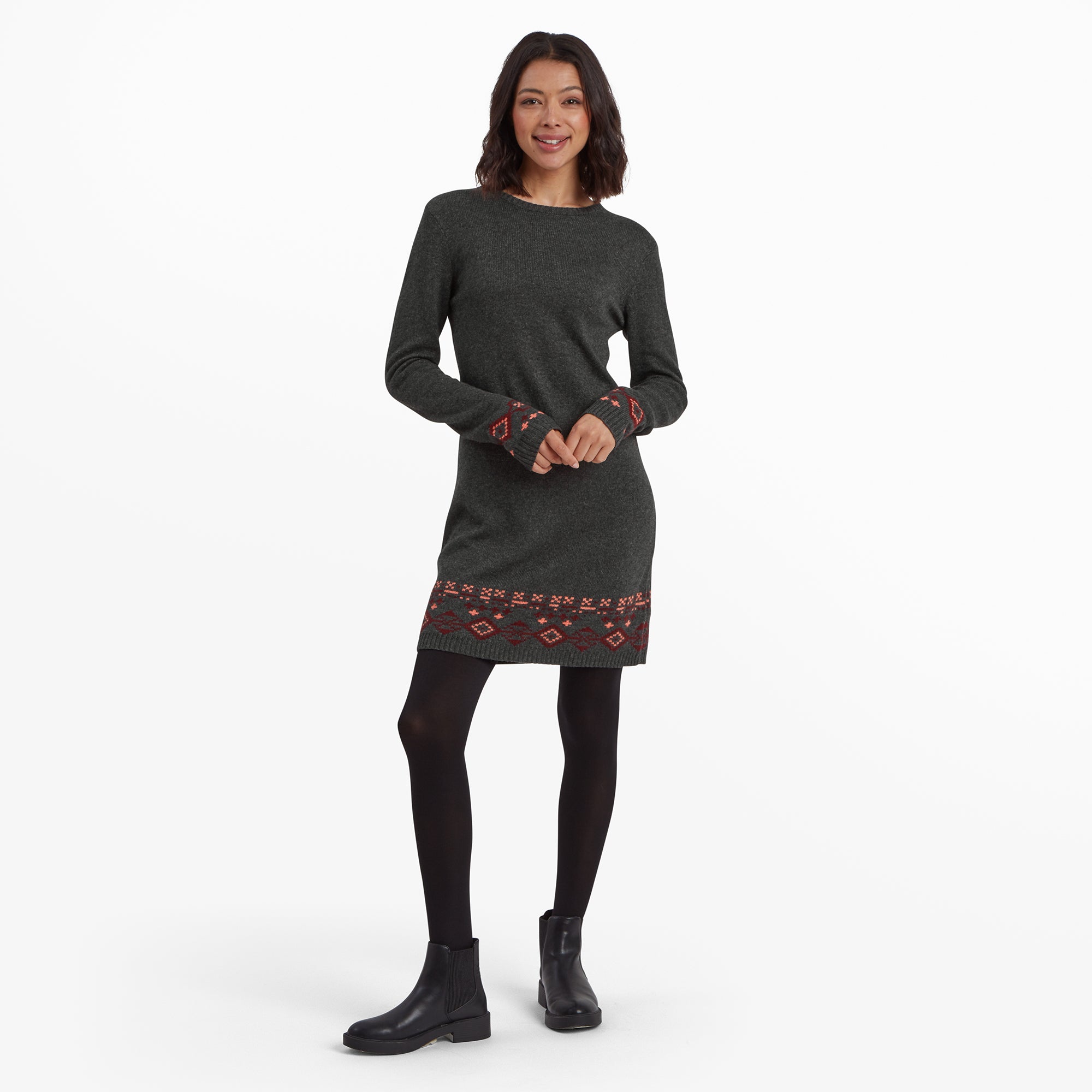 A woman wearing a Sherpa Adventure Gear Bharati Eco Dress in Grey with red and pink geometric patterns at the hem and cuffs. She pairs the outfit with black tights and black Chelsea boots, standing confidently with her arms slightly crossed.