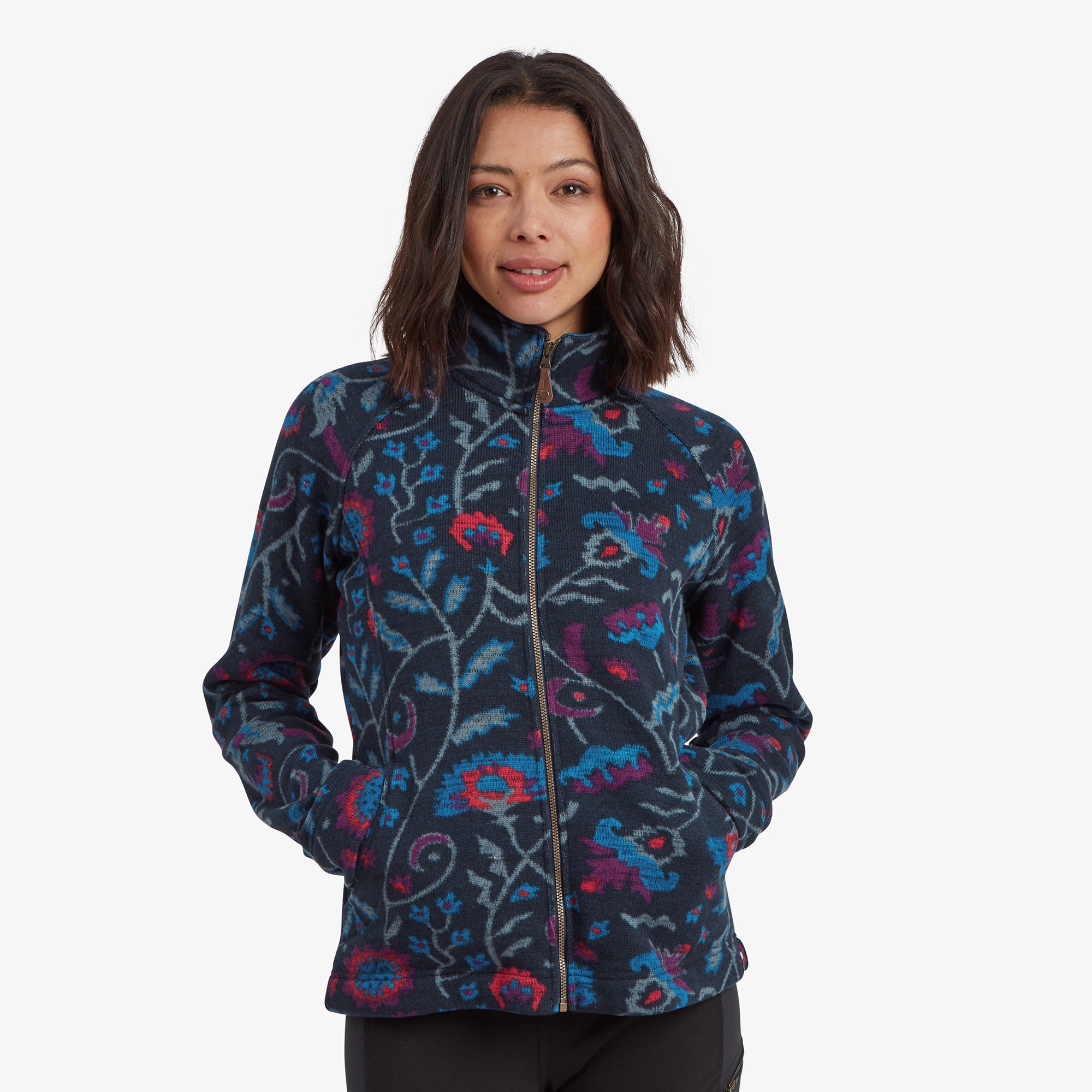 A close-up of a woman wearing a Sherpa Adventure Gear Bhutan Full Zip Jacket in Blue with vibrant floral patterns. The jacket features a high collar and a smooth zipper detail, providing a cozy yet stylish appearance. The model has her hands in the side pockets, showcasing their functionality.