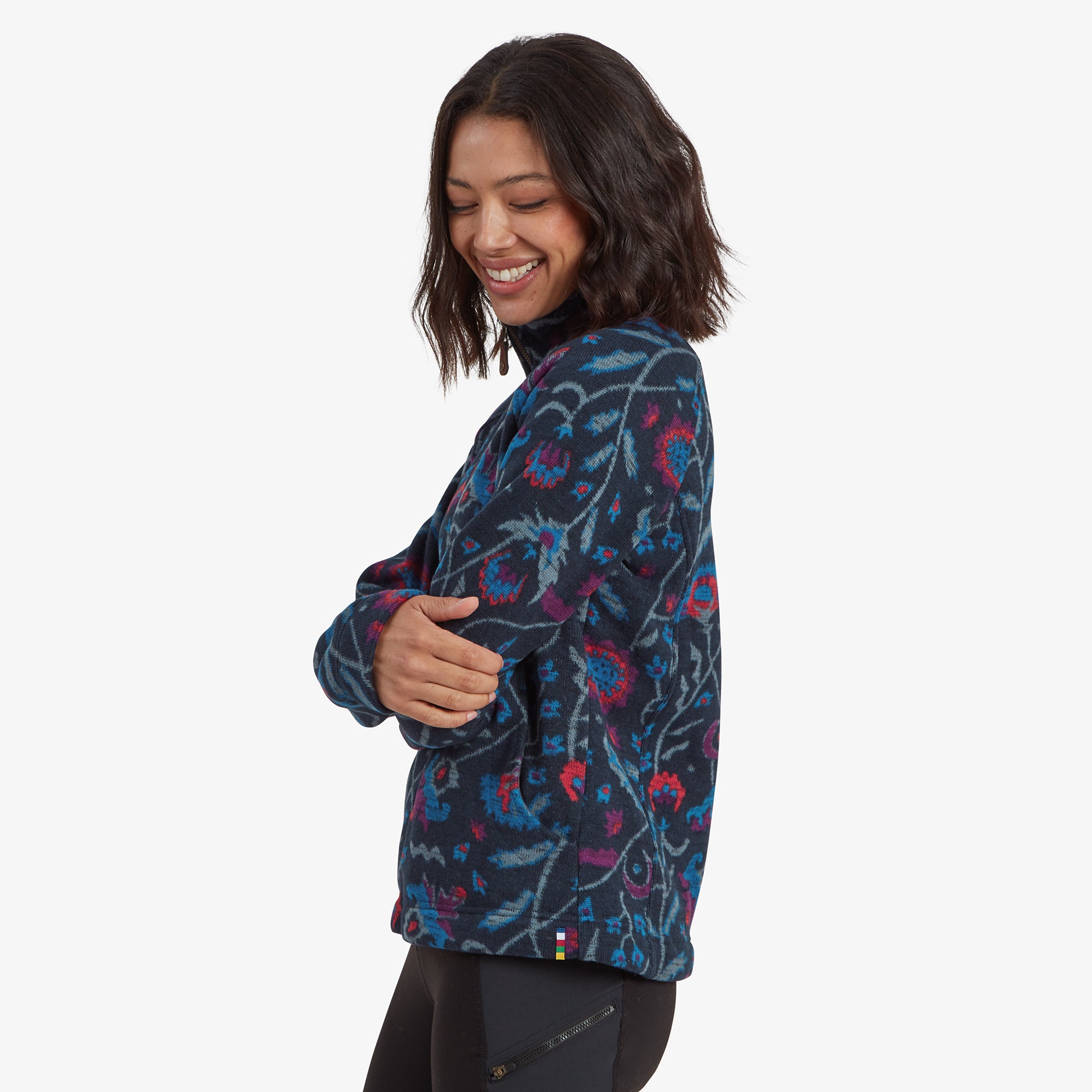 A side view of the Sherpa Adventure Gear Bhutan Full Zip Jacket in Blue, showing its tailored fit and vibrant floral patterns extending to the back. The model smiles gently, emphasising the comfort and stylish cut of the jacket.