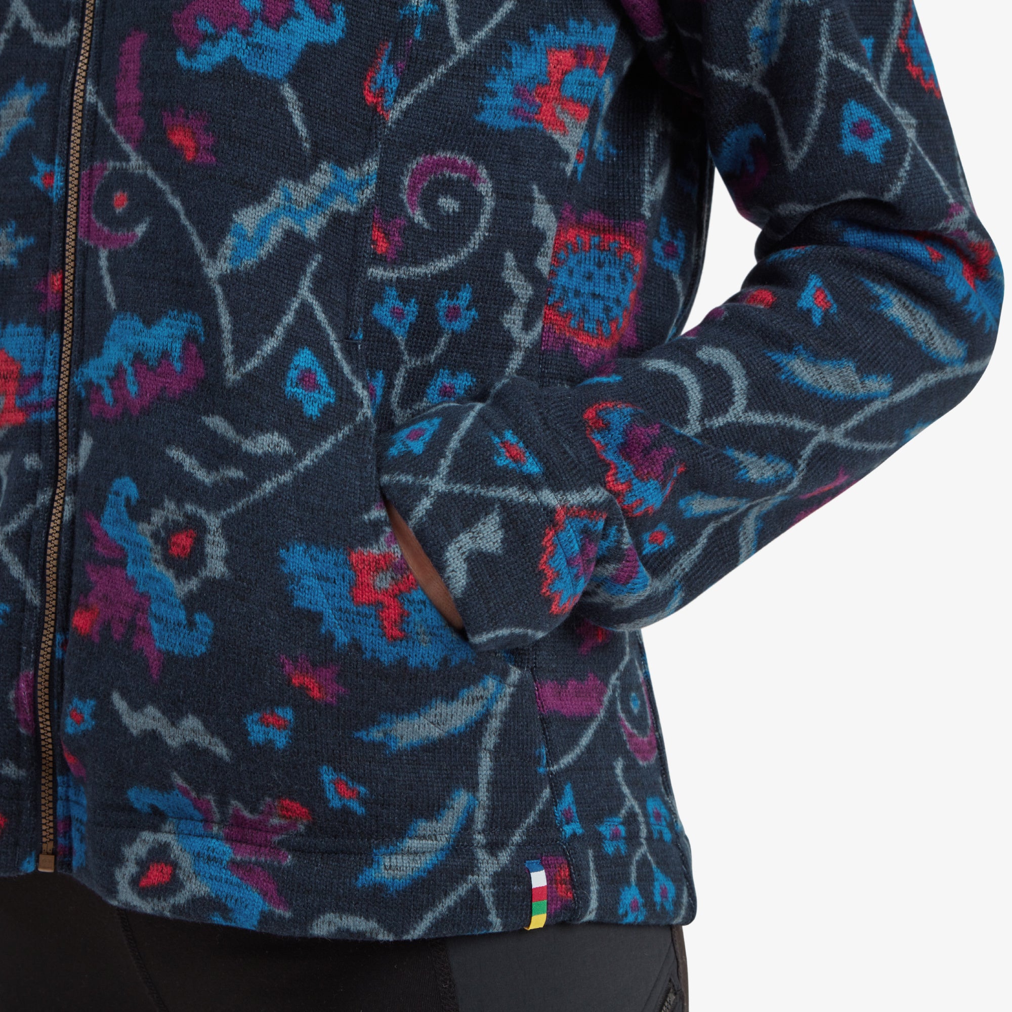 A zoomed-in shot of the side pocket on the Sherpa Adventure Gear Bhutan Full Zip Jacket in Blue, showing its accessibility and the intricate floral detailing around it. A colorful Sherpa flag tag adds a unique and branded touch.