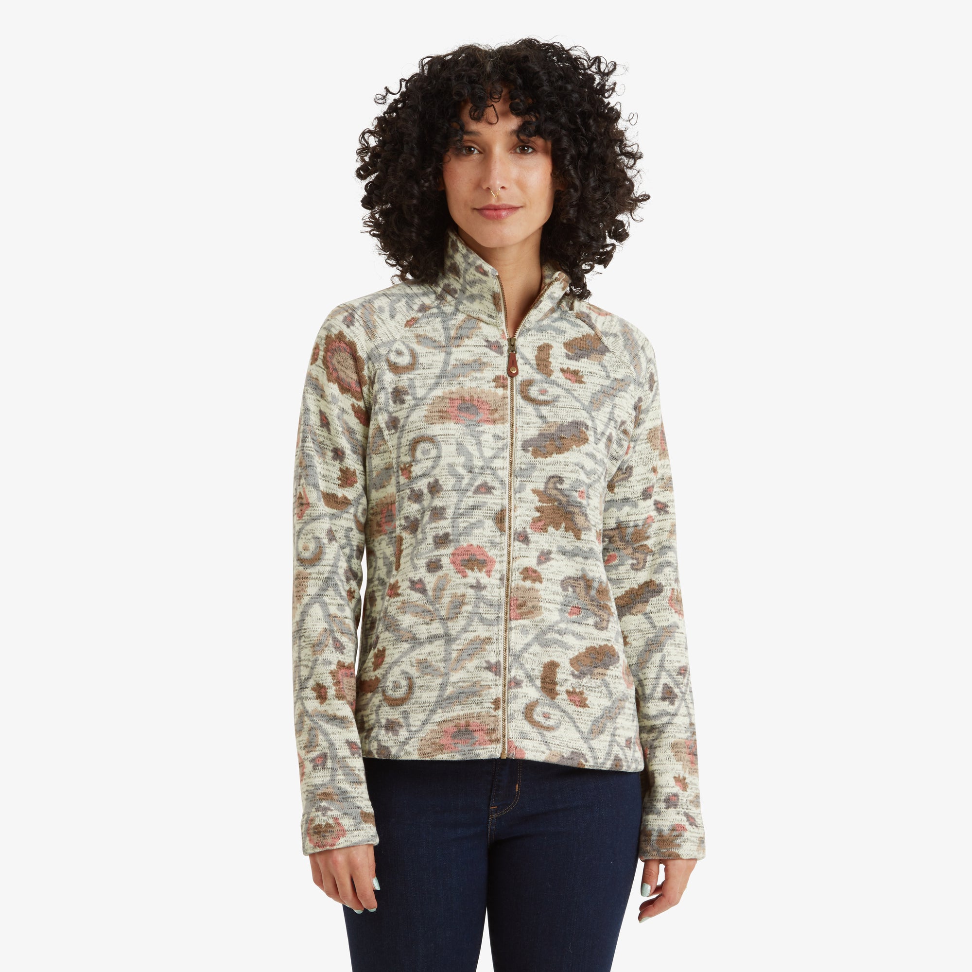 A front-facing image showcasing a woman wearing a Sherpa Adventure Gear Bhutan Full Zip Jacket in Light Brown. The jacket features intricate patterns in earthy tones, a high collar, and a full-zip closure. The model pairs the jacket with dark denim jeans.