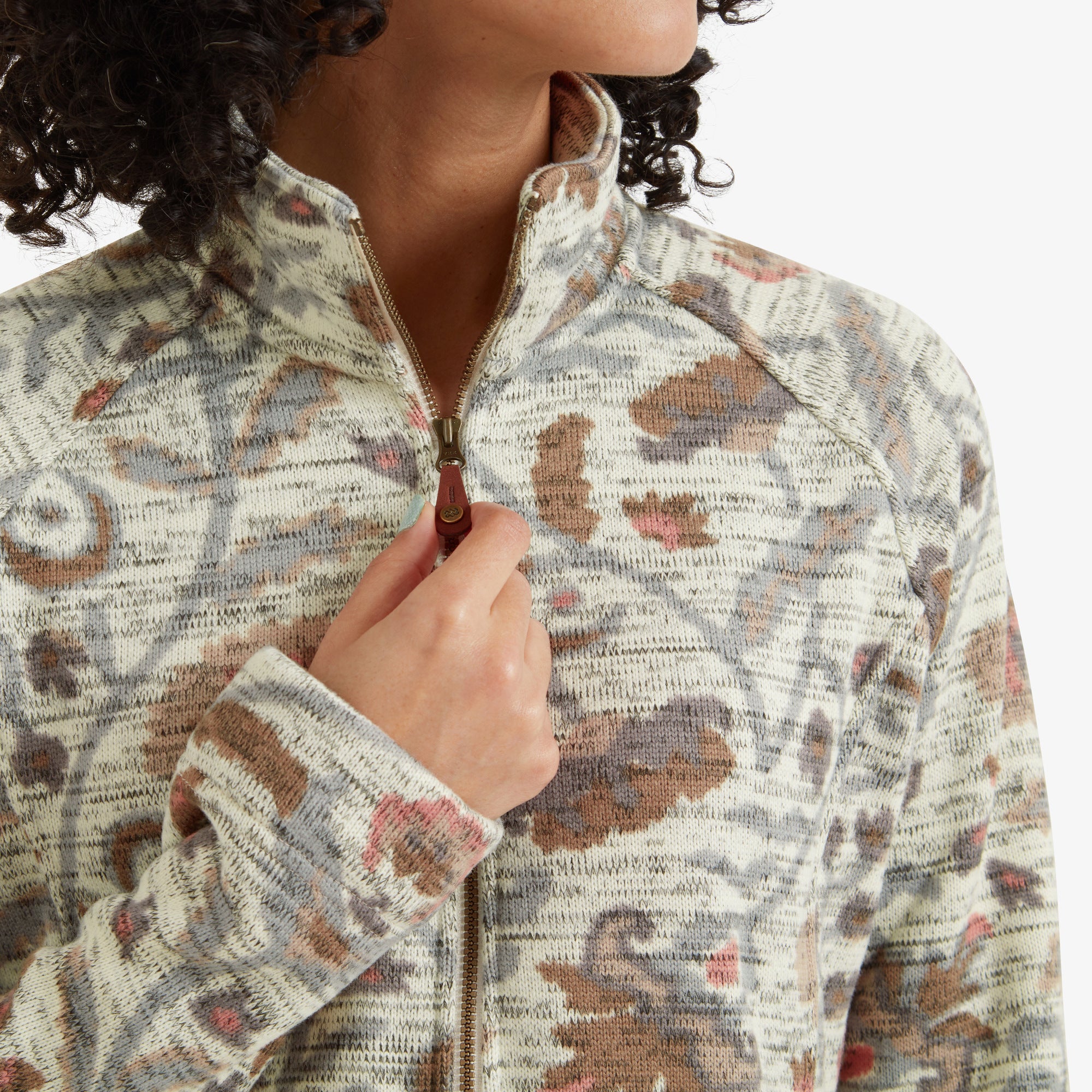 A close-up shot of the model’s hand adjusting the zipper of the Sherpa Adventure Gear Bhutan Full Zip Jacket in Light Brown. The zipper features a leather pull-tab, and the detailed pattern around the collar and chest area is clearly highlighted.