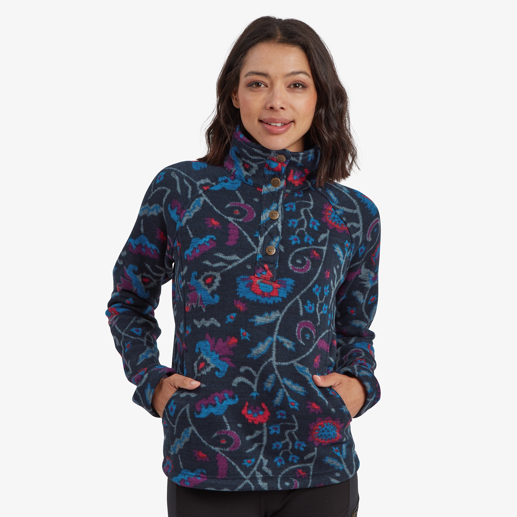 The model wears a vibrant Sherpa Adventure Gear Bhutan Pullover in Blue adorned with a bold floral design in pink, blue, and red tones. The high-neck collar features a button closure with intricate wooden-style buttons, adding a rustic charm. The model's hands rest comfortably in the kangaroo pocket, emphasising its practicality.