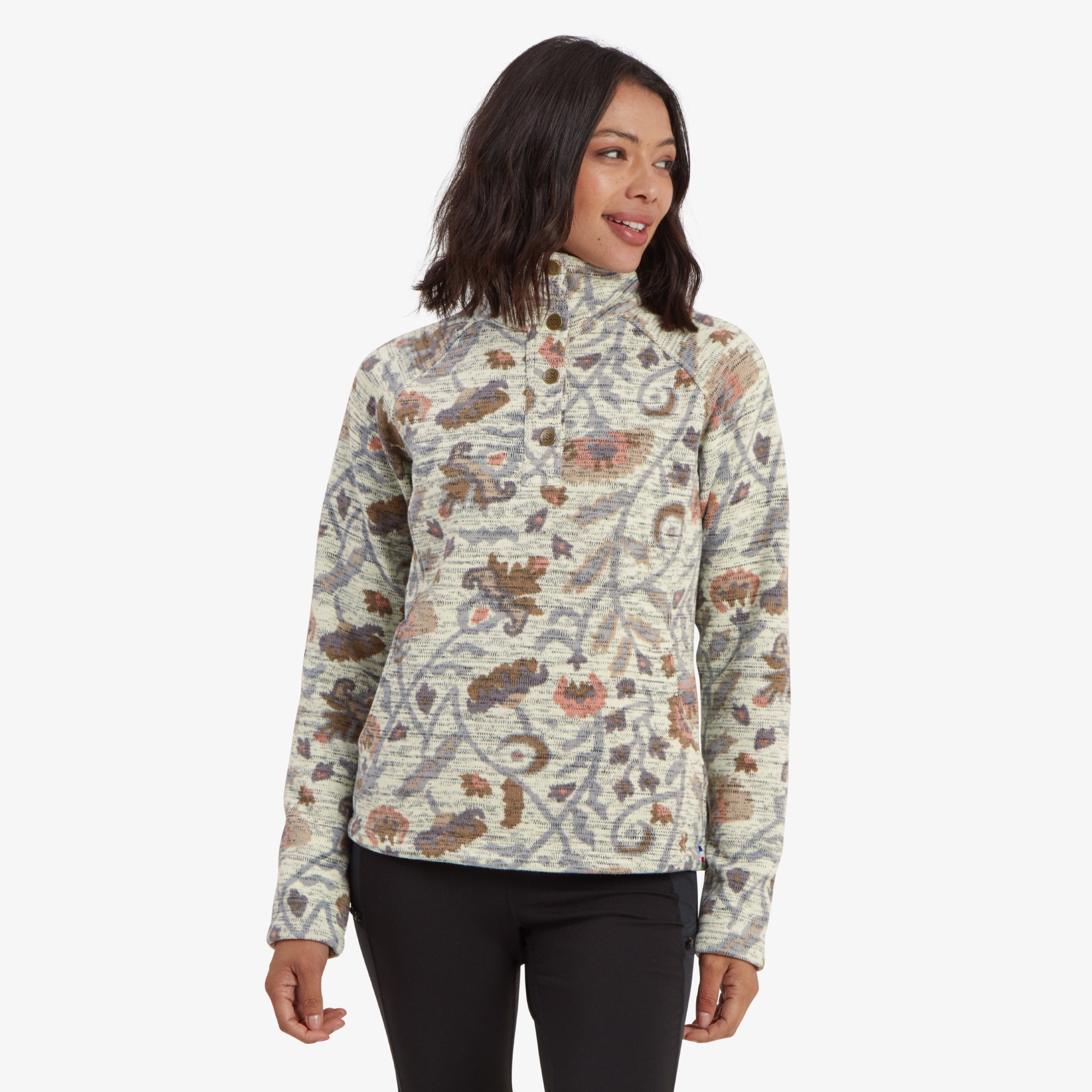 A woman stands facing forward, wearing a Sherpa Adventure Gear Bhutan Pullover in Light Brown with a colorful floral-inspired pattern. The high-neck design features a buttoned placket and long sleeves with thumbholes. The fabric has a textured knit appearance, and the pullover includes discreet hand pockets. She pairs the top with black leggings, showcasing a casual and cozy look.