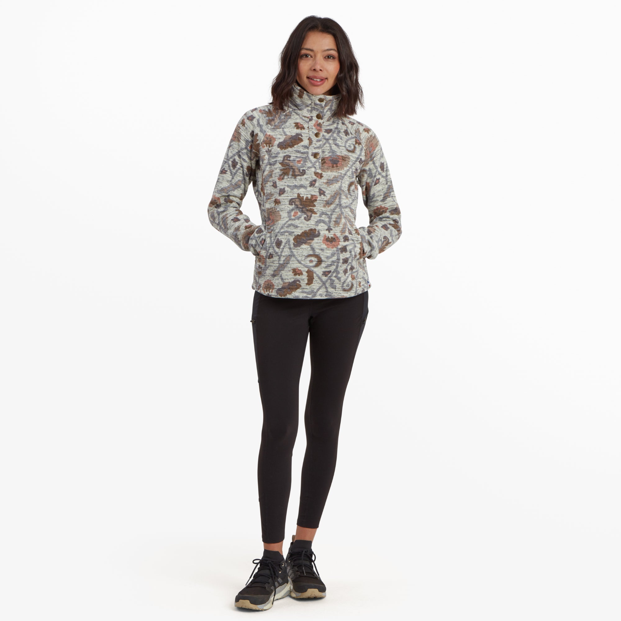 The model stands with her hands in the pockets of the Sherpa Adventure Gear Bhutan Pullover in Light Brown, showcasing its relaxed fit and high-neck design. The floral-inspired motifs in subtle tones of brown, green, and orange complement the pullover's neutral base. She pairs the pullover with black leggings and hiking shoes, emphasising an outdoorsy aesthetic.