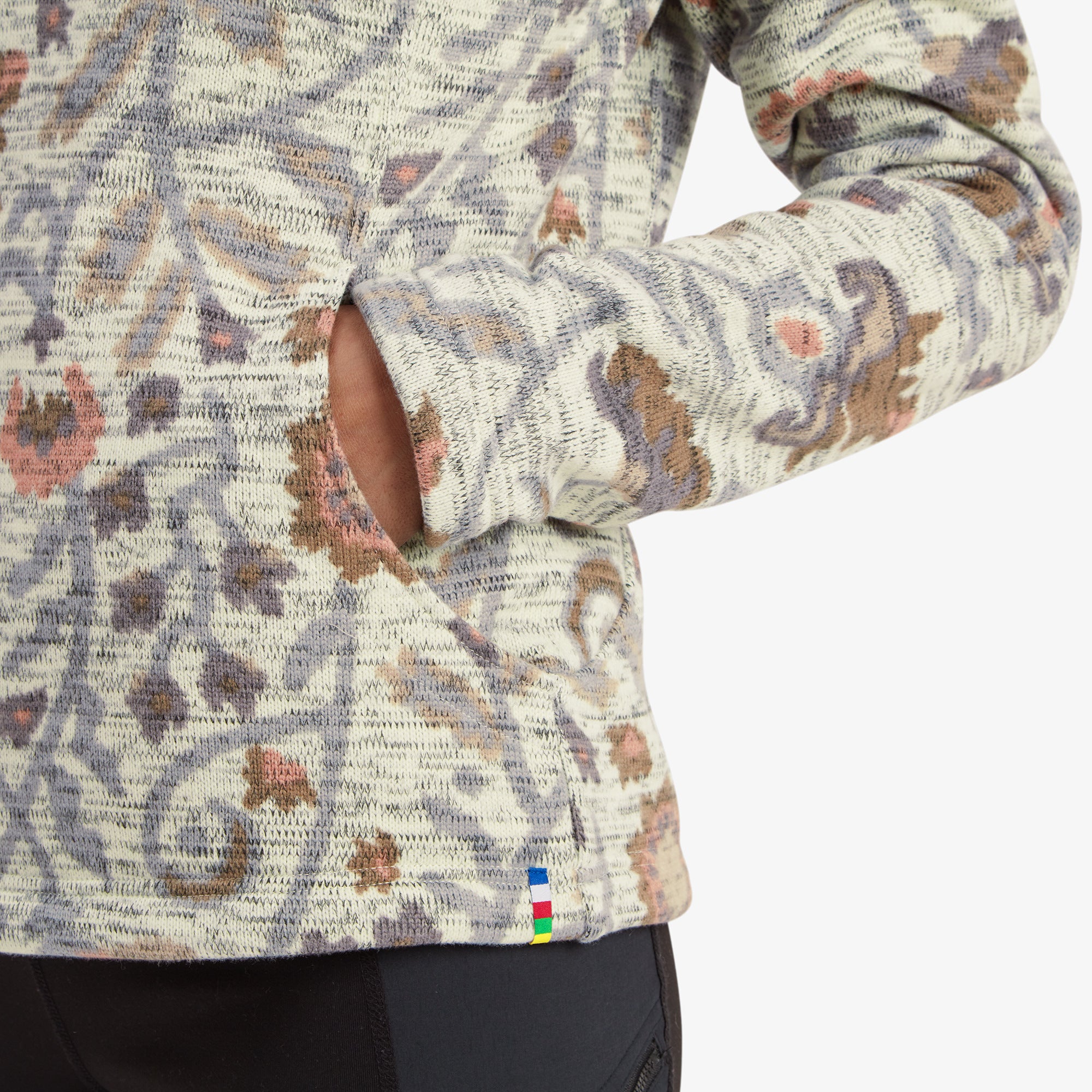 A close-up of the lower part of the Sherpa Adventure Gear Bhutan Pullover in Light Brown shows one hand resting in the side pocket, revealing its functional design. The sleeve features a thumbhole, and the fabric's intricate knit pattern and floral details are highlighted. A small colorful tag at the hem adds a subtle branding touch. The overall texture and craftsmanship are evident in this shot.