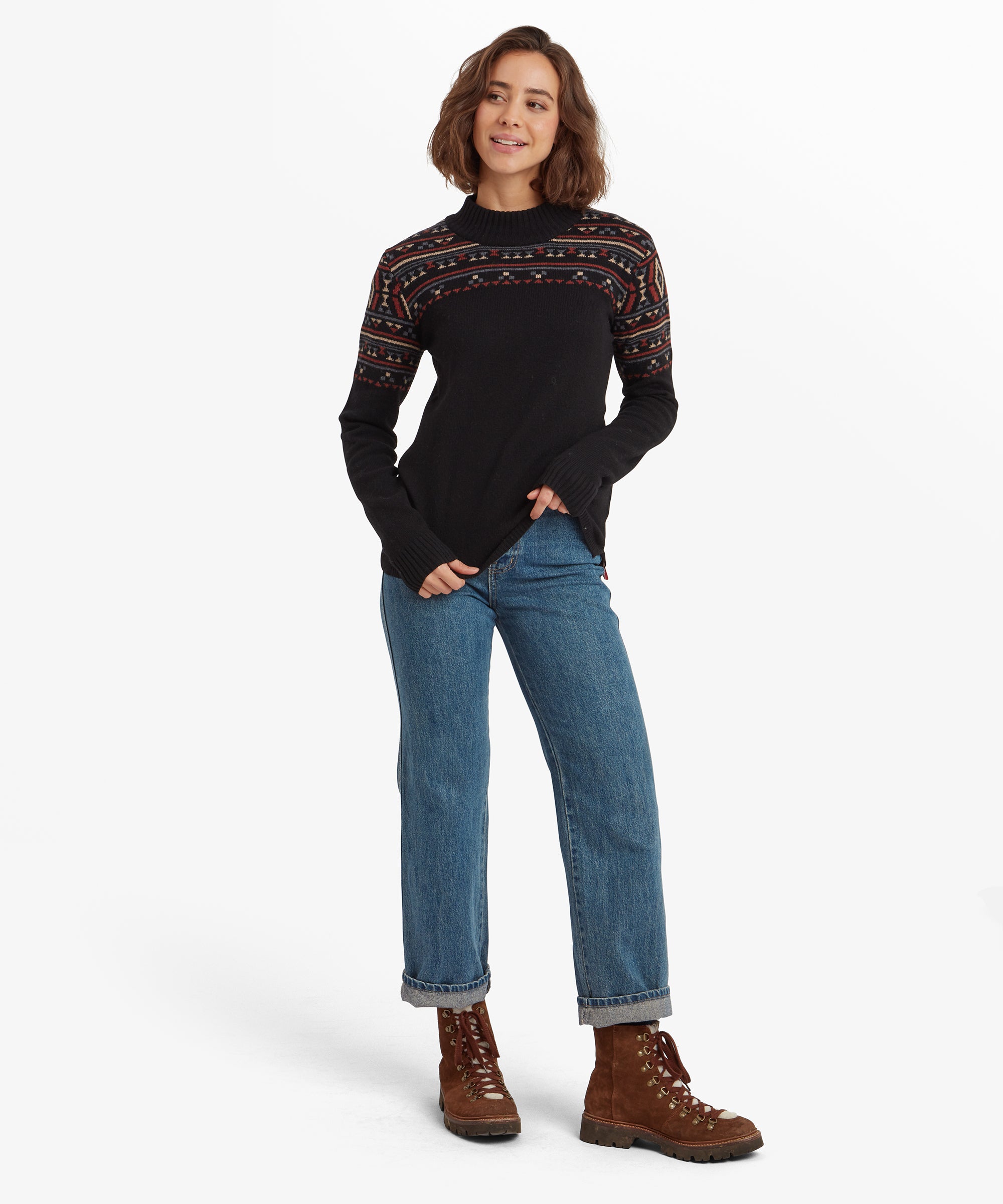 The same woman is shown in a full-body shot, standing with legs apart and hands casually placed at her sides. She is wearing the Sherpa Adventure Gear Bhutla Crew Sweater in Black paired with blue jeans and brown hiking boots. The sweater fits loosely and drapes to her hips.