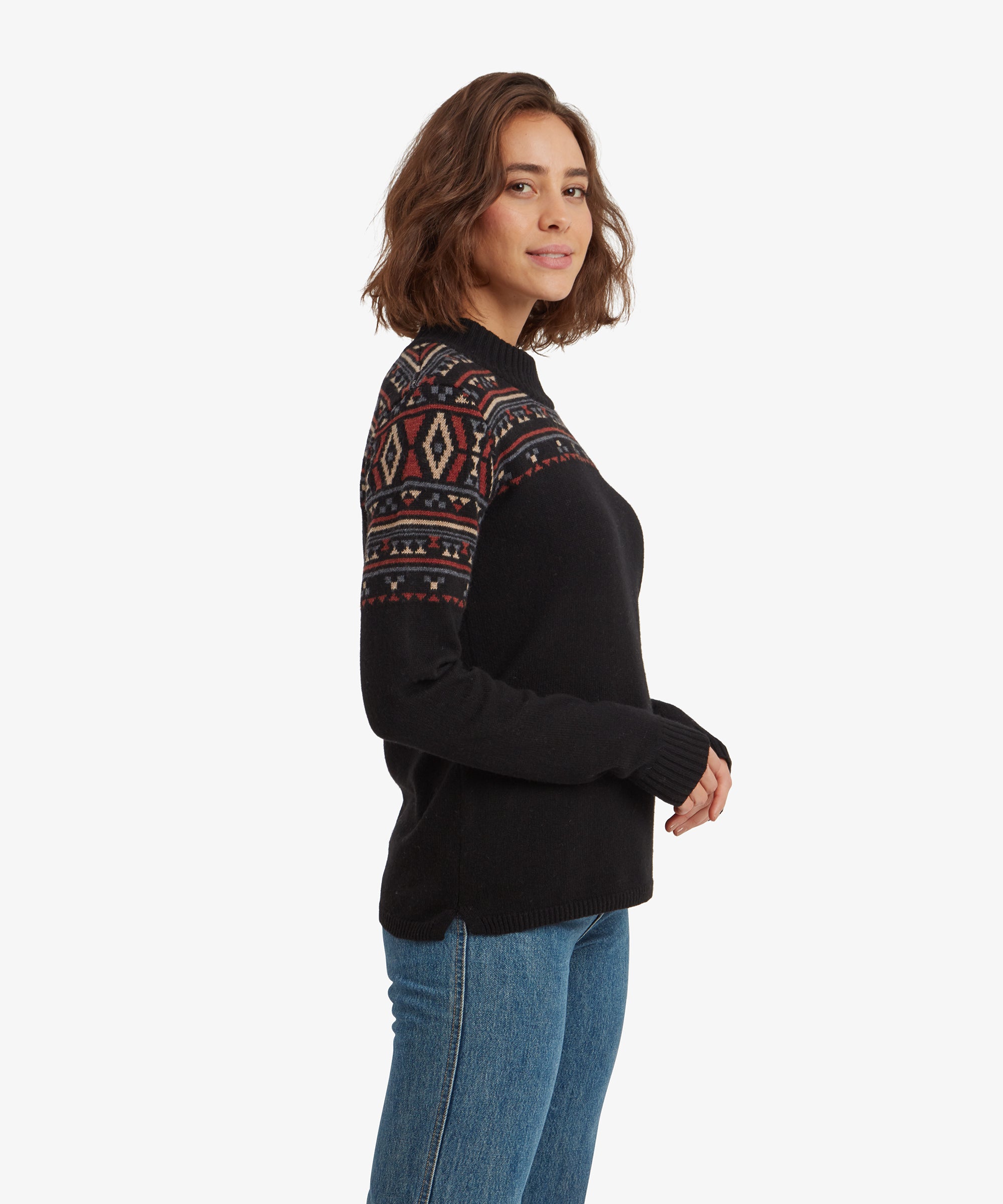 A side profile view of the woman showcases the Sherpa Adventure Gear Bhutla Crew Sweater in Black's fit. The geometric patterns extend from the chest across the shoulders and upper arms. The ribbed neck and hem of the sweater are also visible.