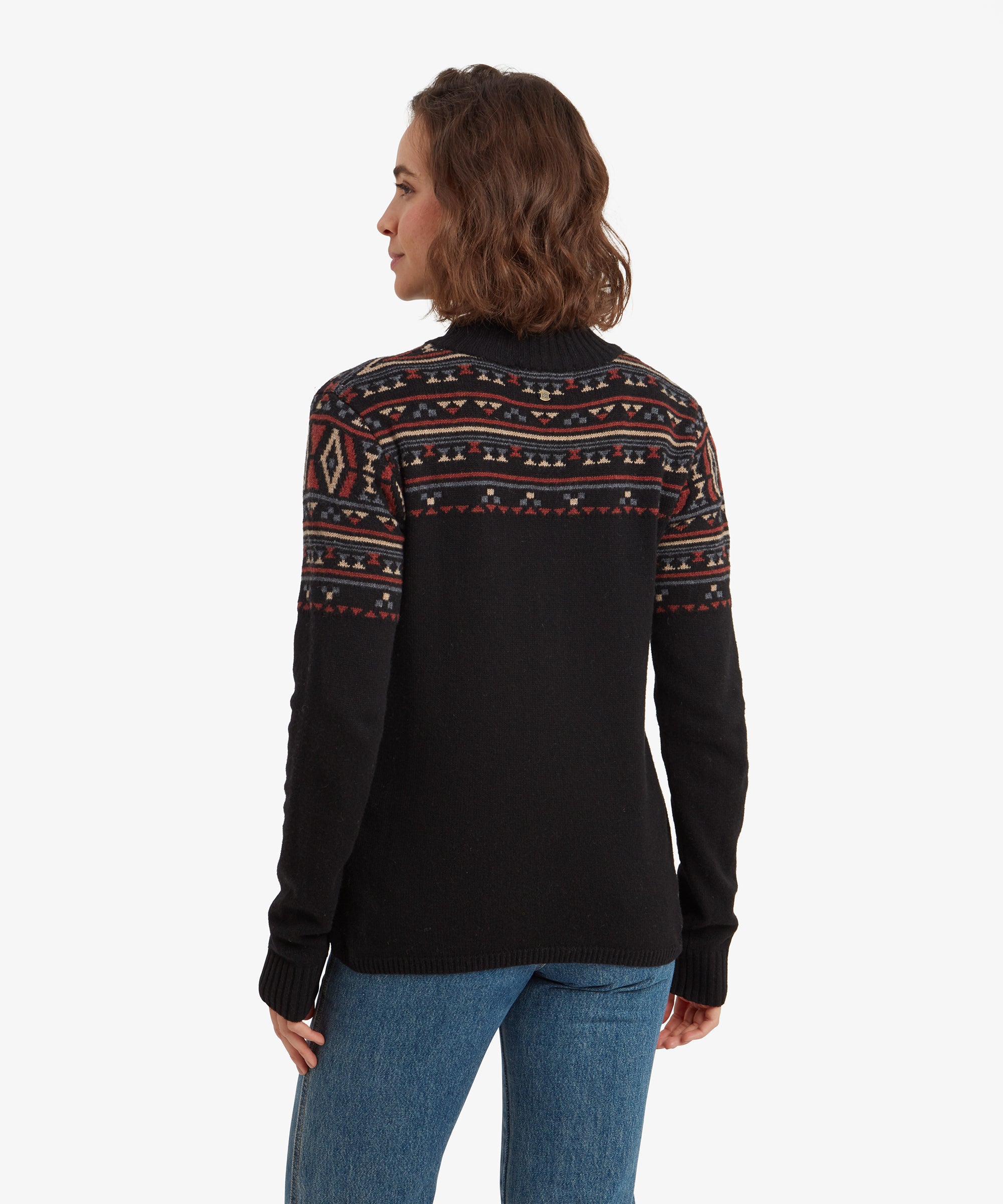 A rear view of the woman wearing the Sherpa Adventure Gear Bhutla Crew Sweater in Black reveals that the geometric patterns continue seamlessly across the back shoulders. The body of the sweater is plain black with a ribbed hem at the bottom.