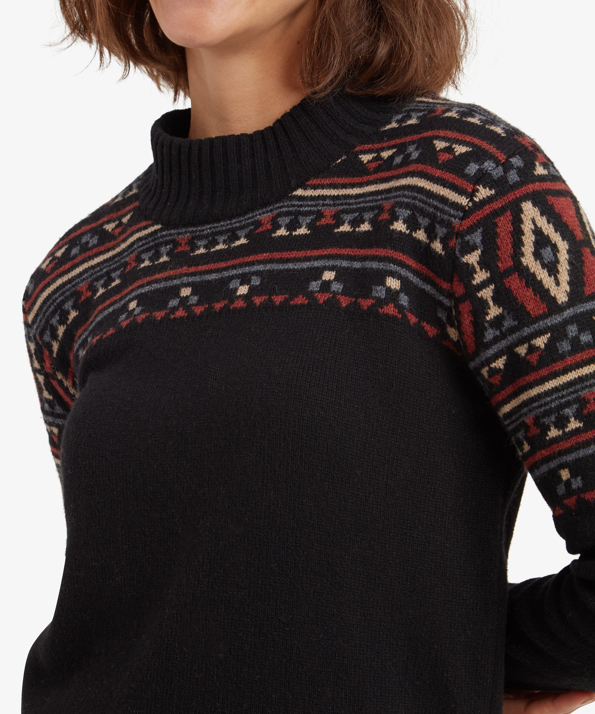 A close-up image of the upper half of the Sherpa Adventure Gear Bhutla Crew Sweater in Black highlights the detailed, multicolored geometric patterns on the shoulders and chest. The ribbed crew neck is prominently featured.