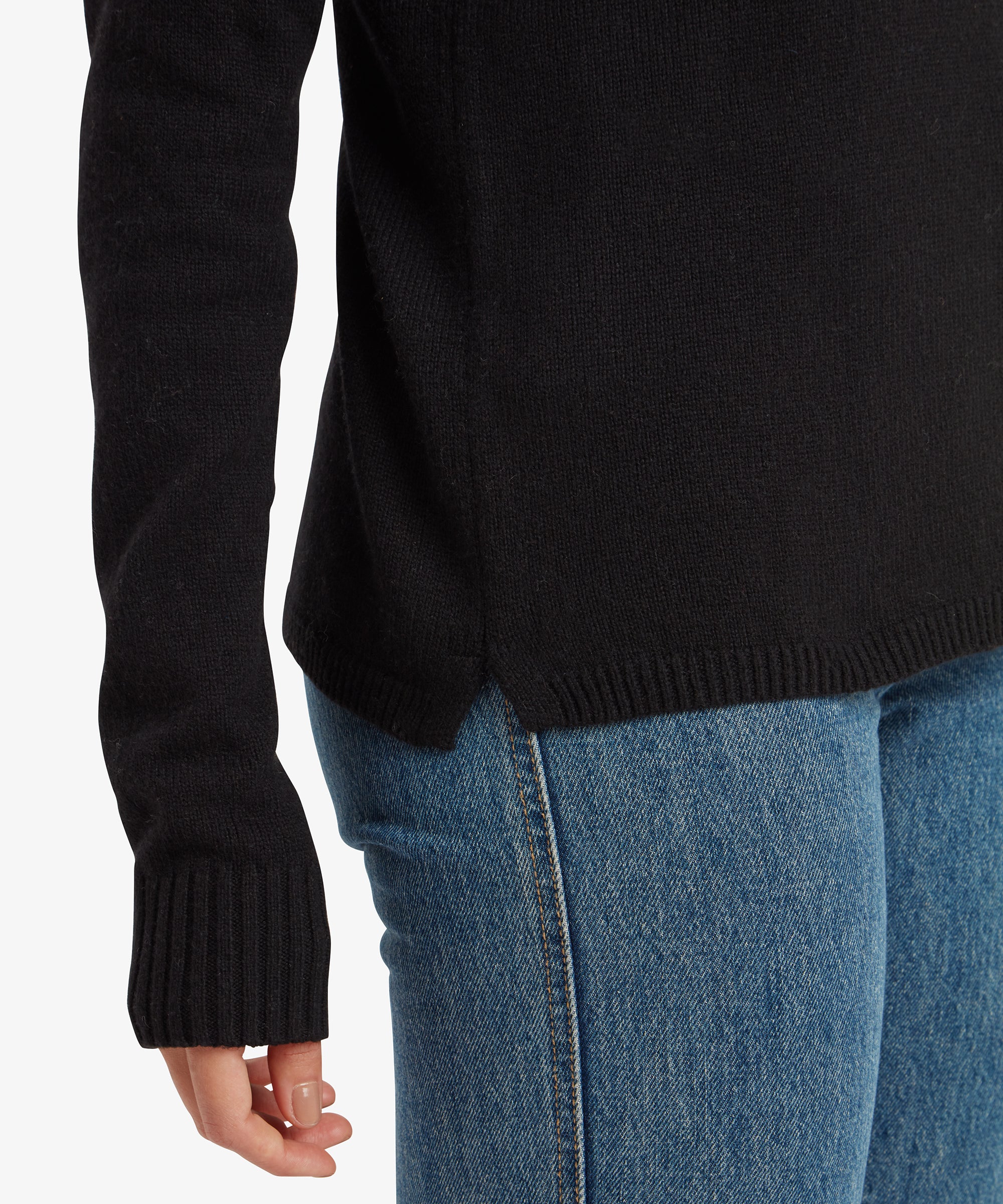 A detailed view of the lower part of the Sherpa Adventure Gear Bhutla Crew Sweater in Black shows the ribbed hem and the long ribbed cuffs extending slightly over the wrist. The woman’s hand is shown relaxed against her blue jeans.