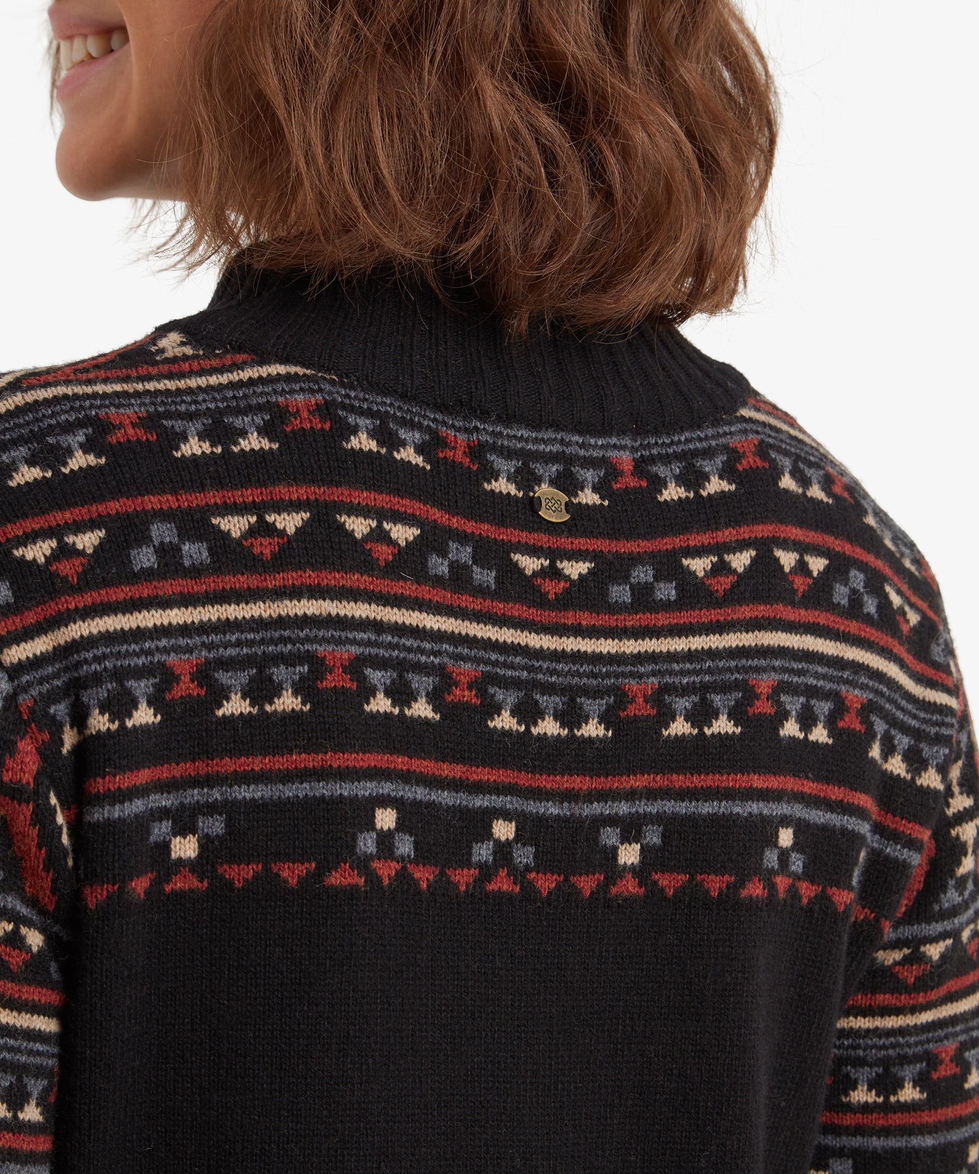 A close-up image of the back neckline showcases the geometric patterns and a small brass logo detail at the center back below the collar. The patterns are symmetrical and incorporate red, beige, and blue hues against the Sherpa Adventure Gear Bhutla Crew Sweater in Black.