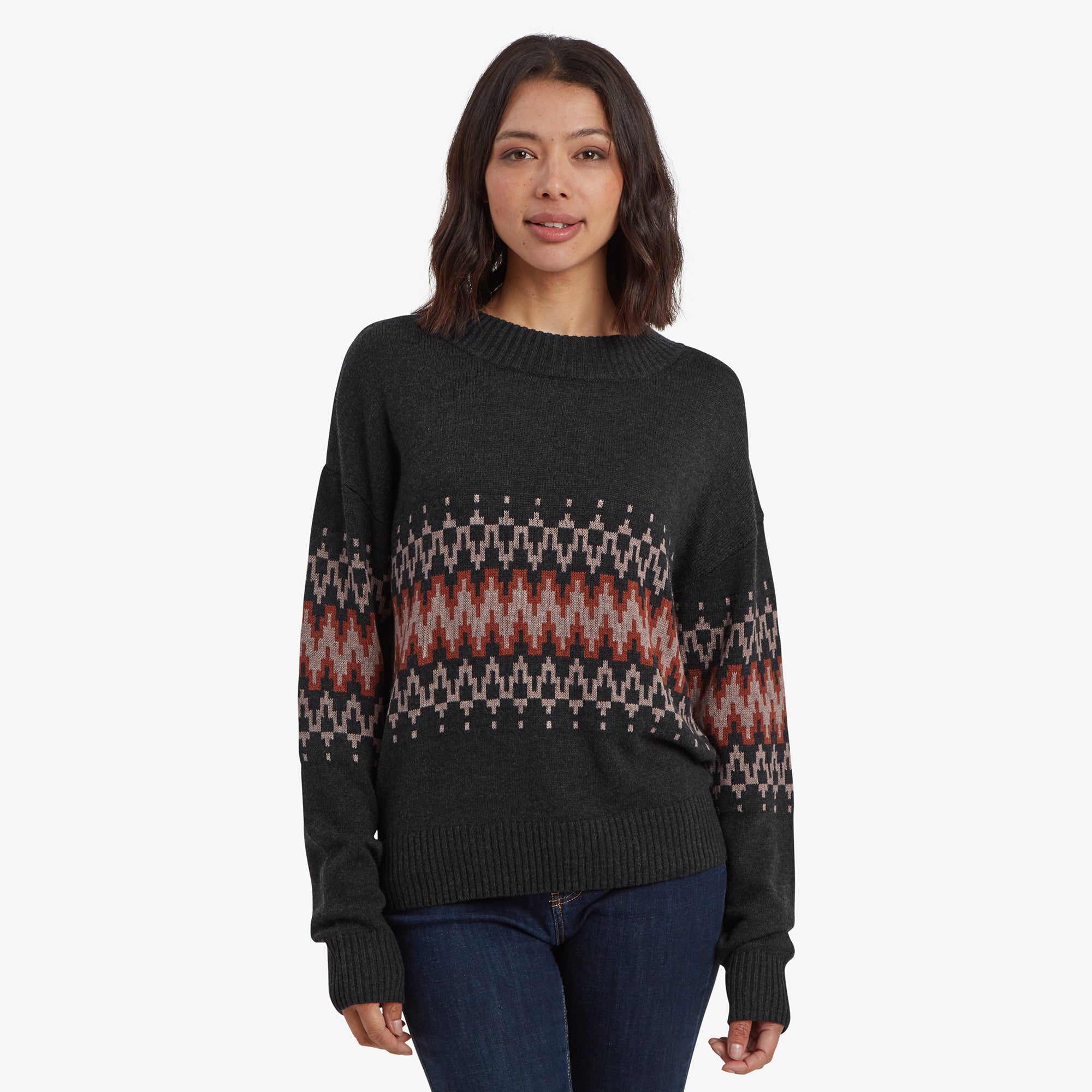 A woman wearing a Sherpa Adventure Gear Devaki Eco Crew in Black with intricate geometric patterns in shades of red, beige, and dark grey across the chest and sleeves. She stands facing the camera, showcasing the relaxed fit and long sleeves of the sweater, paired with dark denim jeans.