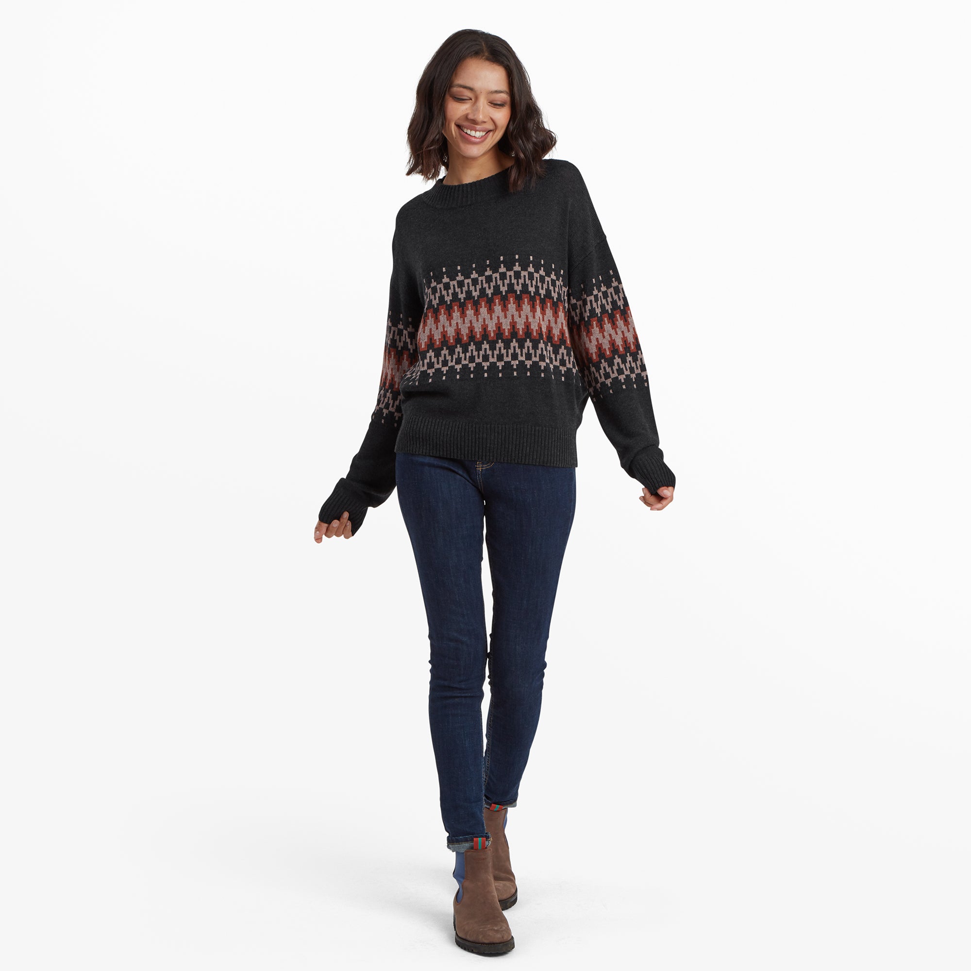 The same woman models the Sherpa Adventure Gear Devaki Eco Crew in Black in a full-body shot, walking with a casual and cheerful demeanor. She wears the sweater with skinny dark jeans and brown ankle boots, providing a complete outfit view and emphasising the comfort and style of the sweater.