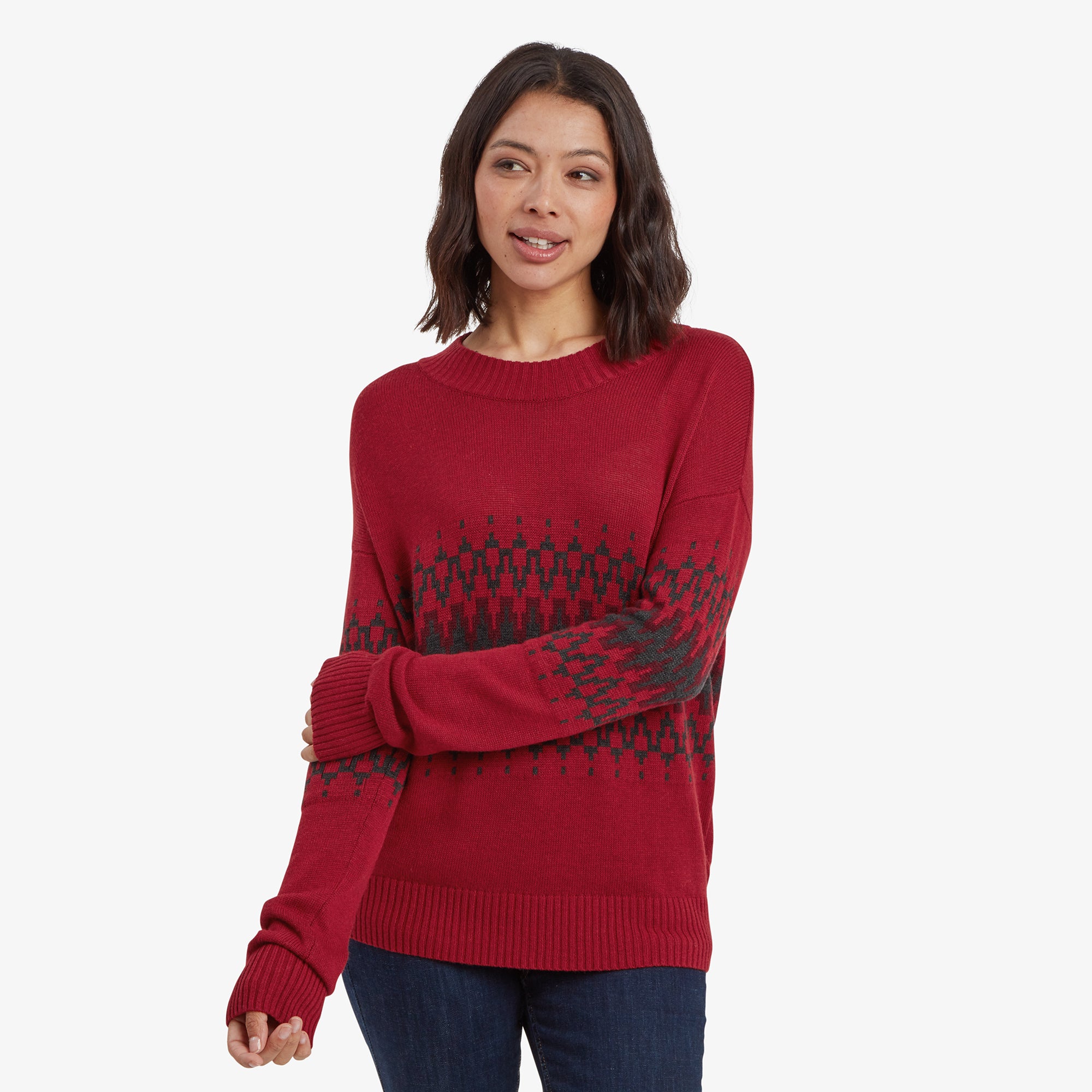 A woman wearing a Sherpa Adventure Gear Devaki Eco Crew Sweater in Red with a crew neckline and a geometric black design pattern across the chest and arms. The sweater has a relaxed fit with ribbed cuffs and hem, paired with dark blue jeans. She is standing in a casual pose with her arms slightly crossed and a soft smile.