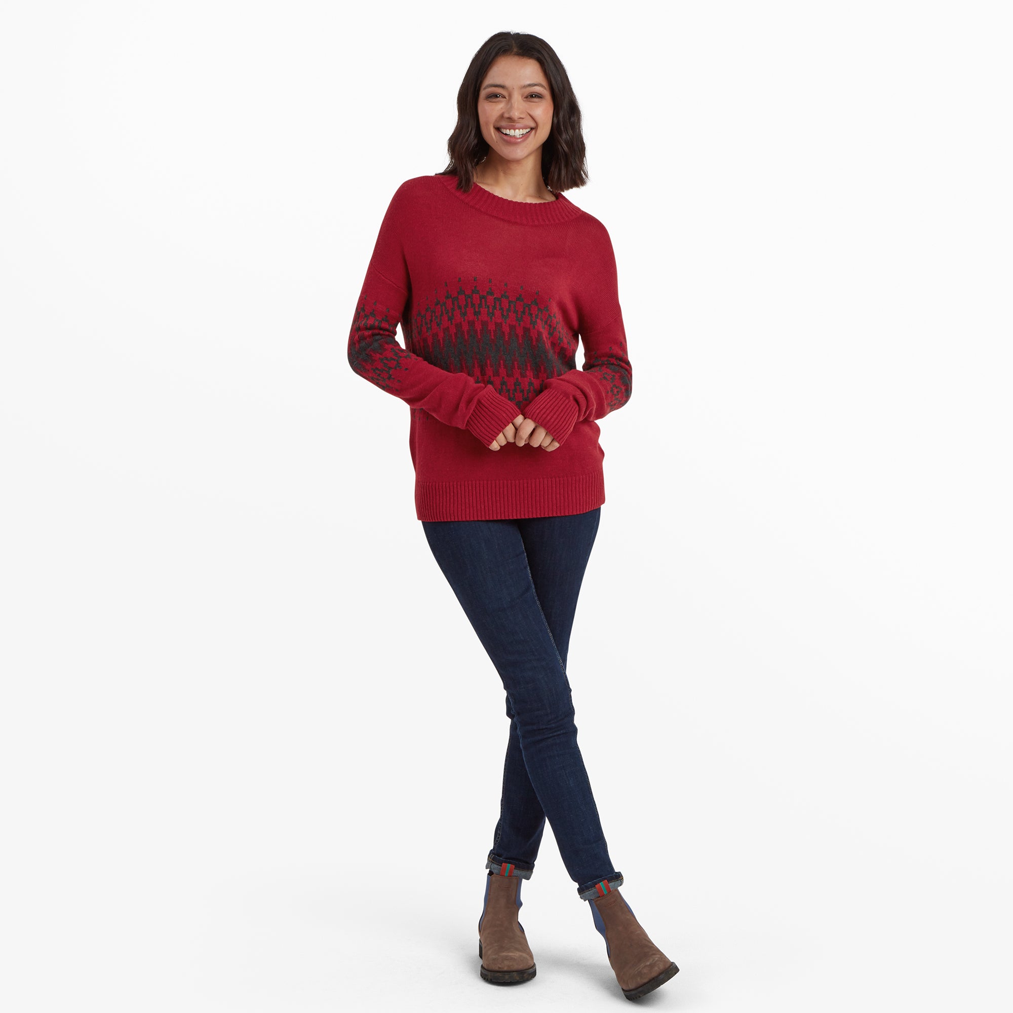 The same woman is captured in a full-body shot, wearing the Sherpa Adventure Gear Devaki Eco Crew Sweater in Red with the black geometric pattern. She pairs it with dark blue jeans and brown ankle boots with colorful striped accents. She is smiling warmly with her hands clasped in front of her, standing confidently.