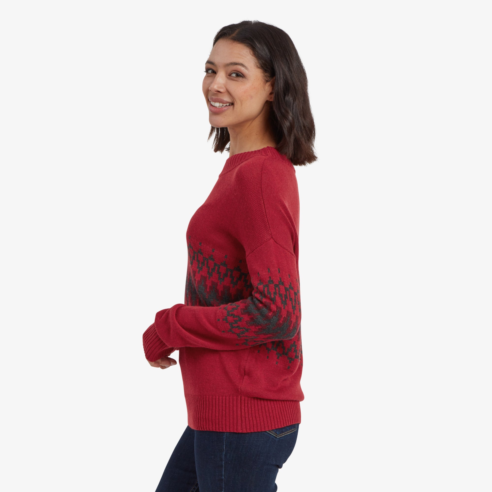 A side profile of the woman in the Sherpa Adventure Gear Devaki Eco Crew Sweater in Red, showcasing the fit of the garment on her torso and sleeves. She is smiling while looking over her shoulder, highlighting the sweater's relaxed yet flattering fit.