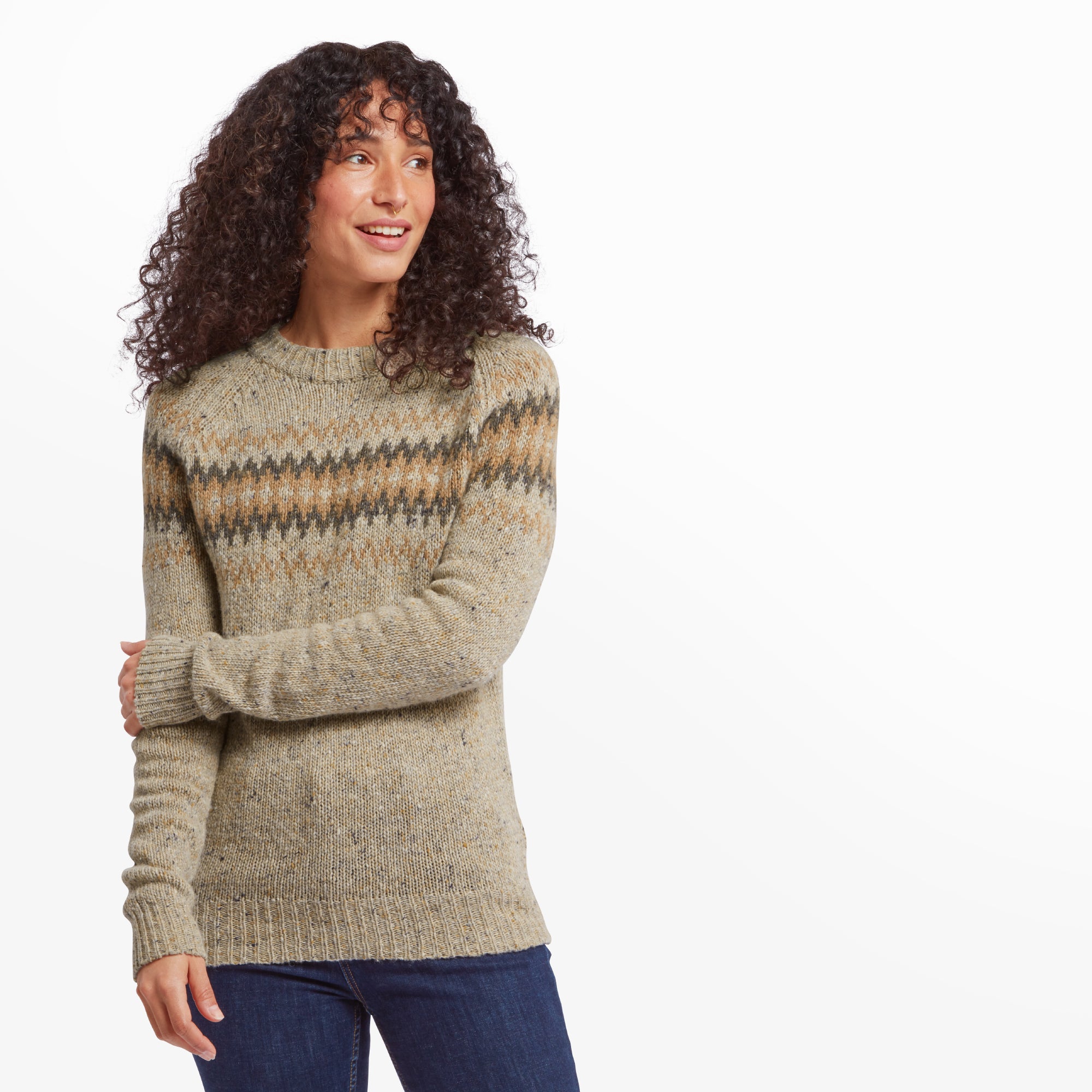 A woman with long curly hair wearing a Sherpa Adventure Gear Dumji Crew Sweater in Beige with an intricate, colorful chevron pattern across the chest. She is smiling softly, standing with one arm crossed over her chest. The sweater features a chunky knit texture and a ribbed hem, cuffs, and neckline. She pairs the sweater with dark blue jeans for a casual, cozy look.