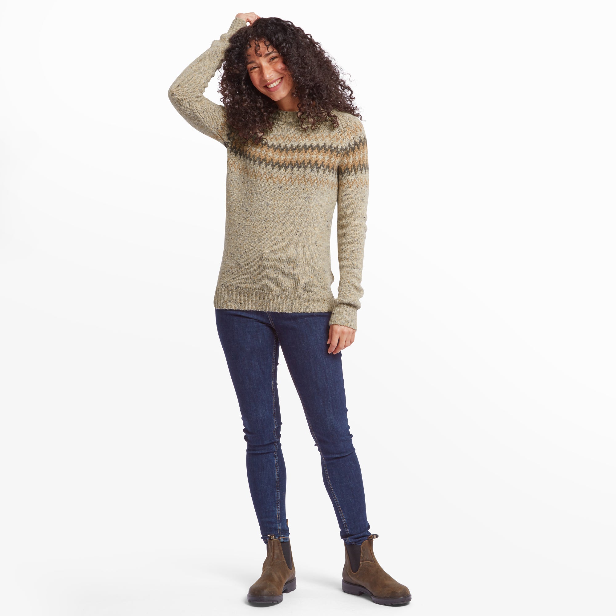 The same woman in the Sherpa Adventure Gear Dumji Crew Sweater in Beige stands with a playful pose, her left arm bent as she touches her head, and her right arm relaxed at her side. She smiles warmly, showcasing the sweater's relaxed fit and soft texture. She wears dark blue skinny jeans and brown Chelsea boots, completing a stylish and practical outfit.