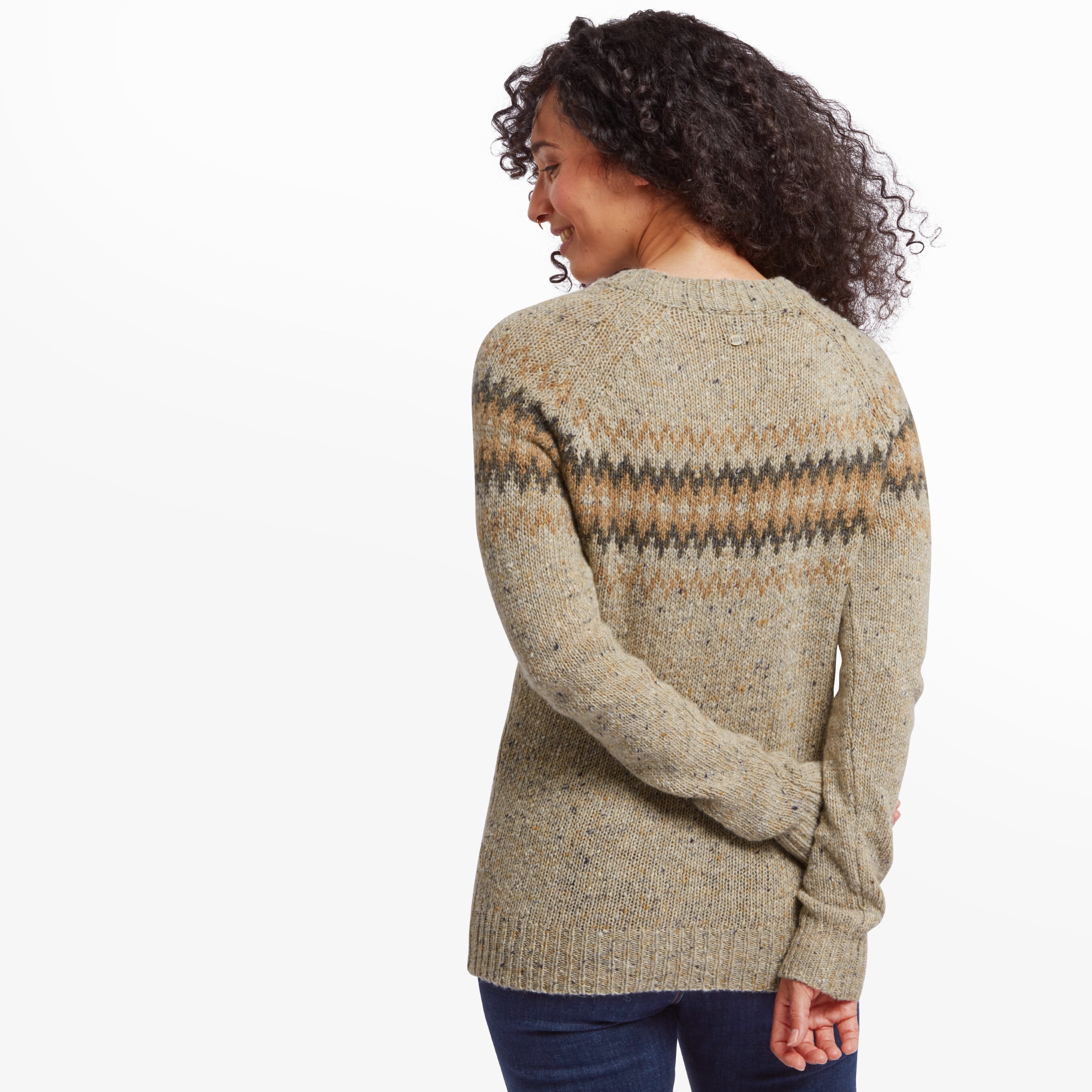 A rear view of the woman in the Sherpa Adventure Gear Dumji Crew Sweater in Beige, highlighting the chevron pattern that extends to the back. She holds her arms behind her, emphasising the slightly fitted silhouette of the sweater. Her curly hair cascades down her back, complementing the cozy, casual aesthetic.