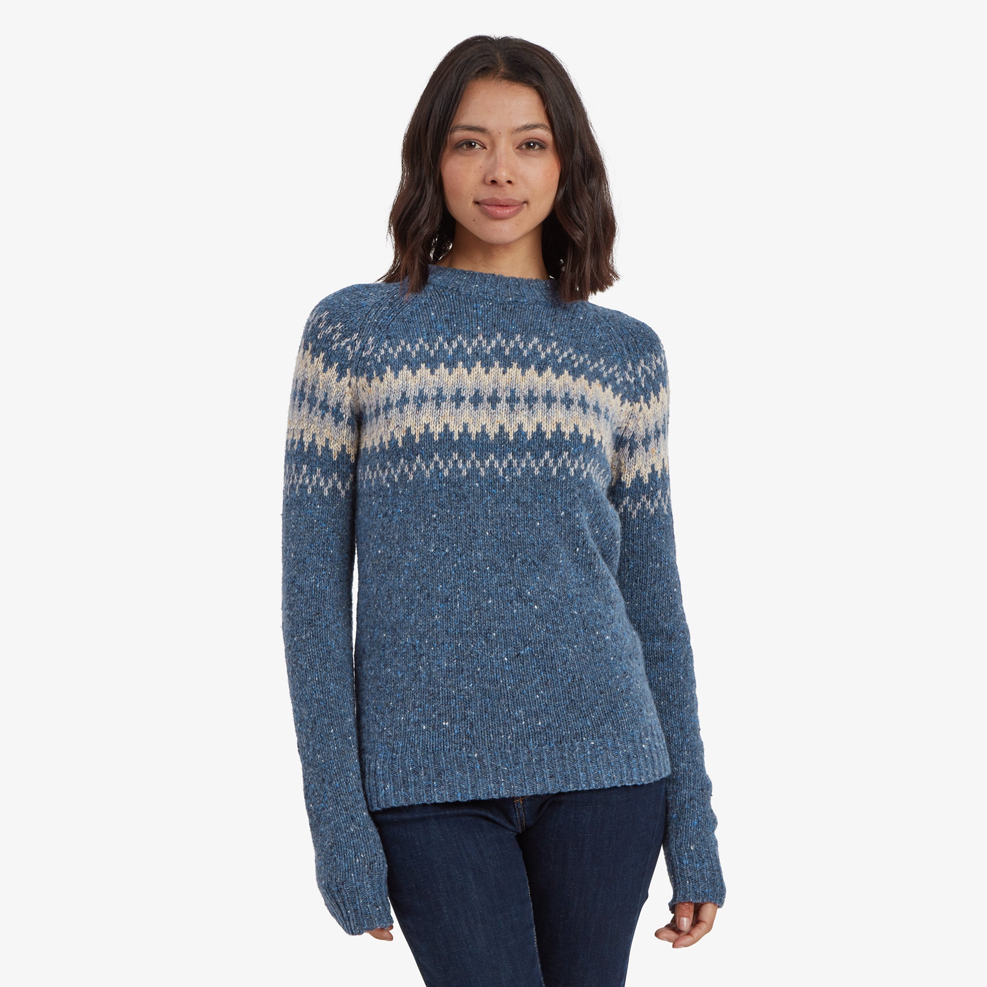 A female model wearing a Sherpa Adventure Gear Dumji Crew Sweater in Blue with beige and light gray Nordic-style patterning across the chest and shoulders. The sweater has a cozy knit texture, long sleeves, and a slightly relaxed fit. The model is facing forward with a neutral expression, showcasing the overall design of the sweater.