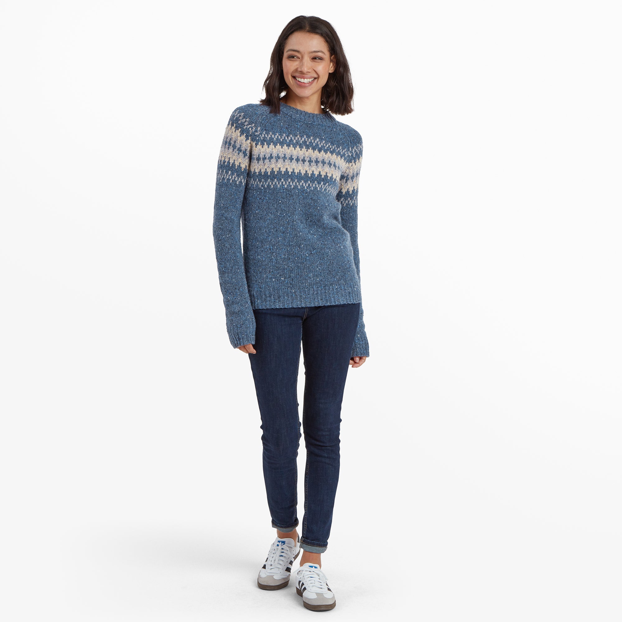 The same model wearing the Sherpa Adventure Gear Dumji Crew Sweater in Blue paired with dark blue skinny jeans and white trainers. She is standing confidently, smiling, with her hands relaxed by her sides. The sweater's length and comfortable fit are visible in this full-body shot, highlighting its casual and versatile look.