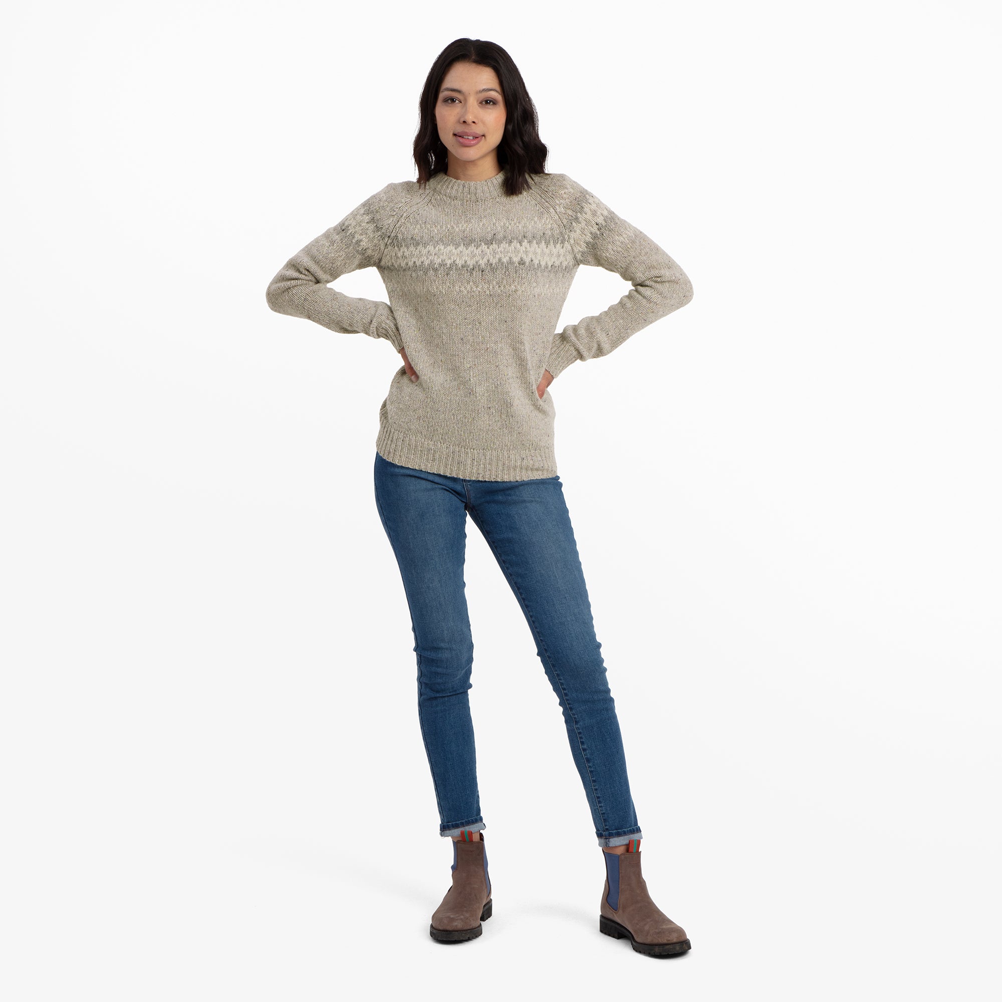 The model is posing confidently, wearing the Sherpa Adventure Gear Dumji Crew Sweater in Brown with a zigzag design. Her hands rest on her hips, and she is paired with fitted blue jeans and brown ankle boots with blue elastic inserts. The background is white, highlighting the outfit.