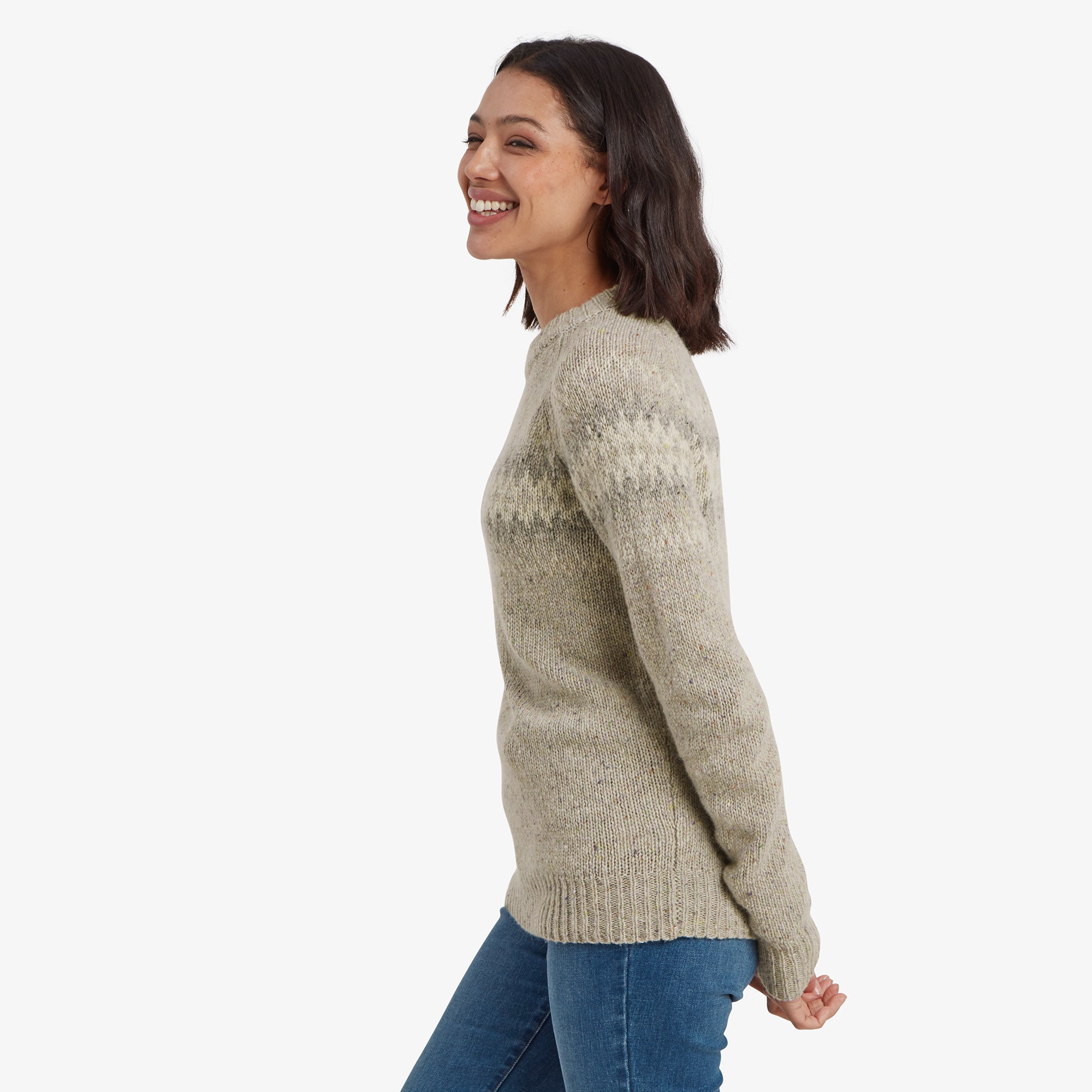 The model is captured in a side profile, smiling brightly. She is wearing the same Sherpa Adventure Gear Dumji Crew Sweater in Brown and blue jeans. The zigzag pattern is visible from the side, emphasising the texture and detailing of the sweater. Her relaxed pose adds a casual feel to the look.