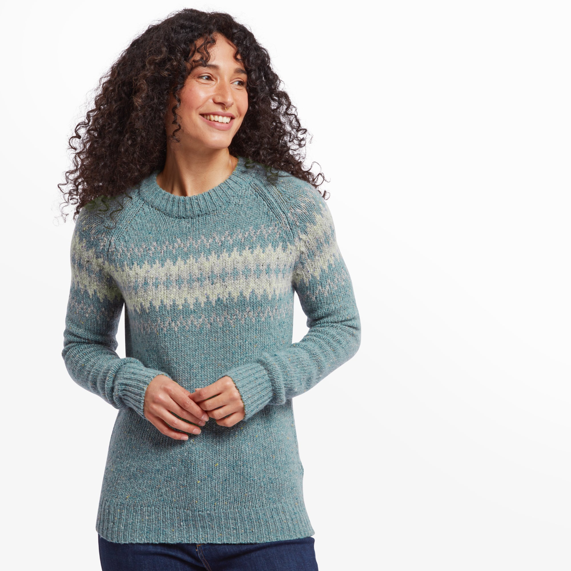 A smiling woman wearing a Sherpa Adventure Gear Dumji Crew Sweater in Green. The sweater features a classic crew neck and a fair isle-inspired pattern across the chest in subtle shades of white and beige. She has natural curly hair and is standing against a white background, wearing dark blue jeans.