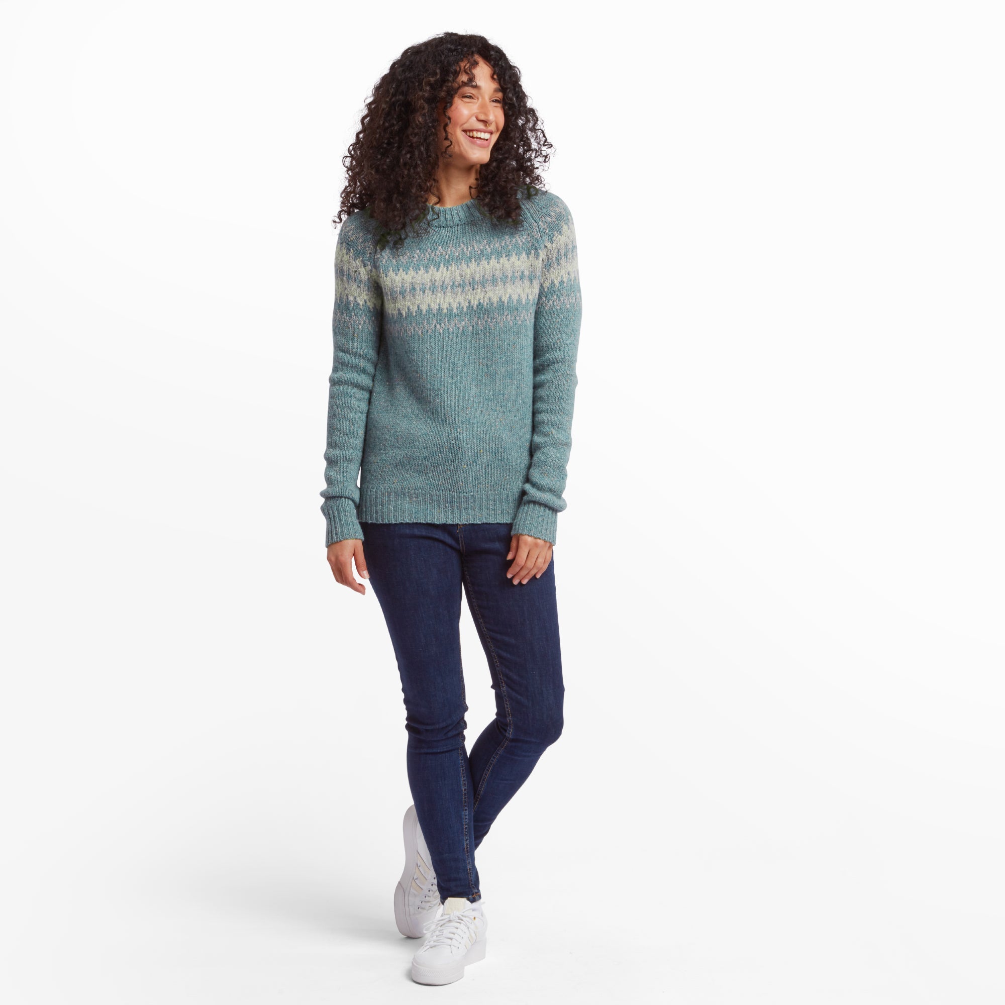 Full-body image of a woman wearing a Sherpa Adventure Gear Dumji Crew Sweater in Green with a fair isle pattern. She pairs it with dark blue skinny jeans and white trainers, standing casually with one leg slightly bent. The background is a clean white studio setting.