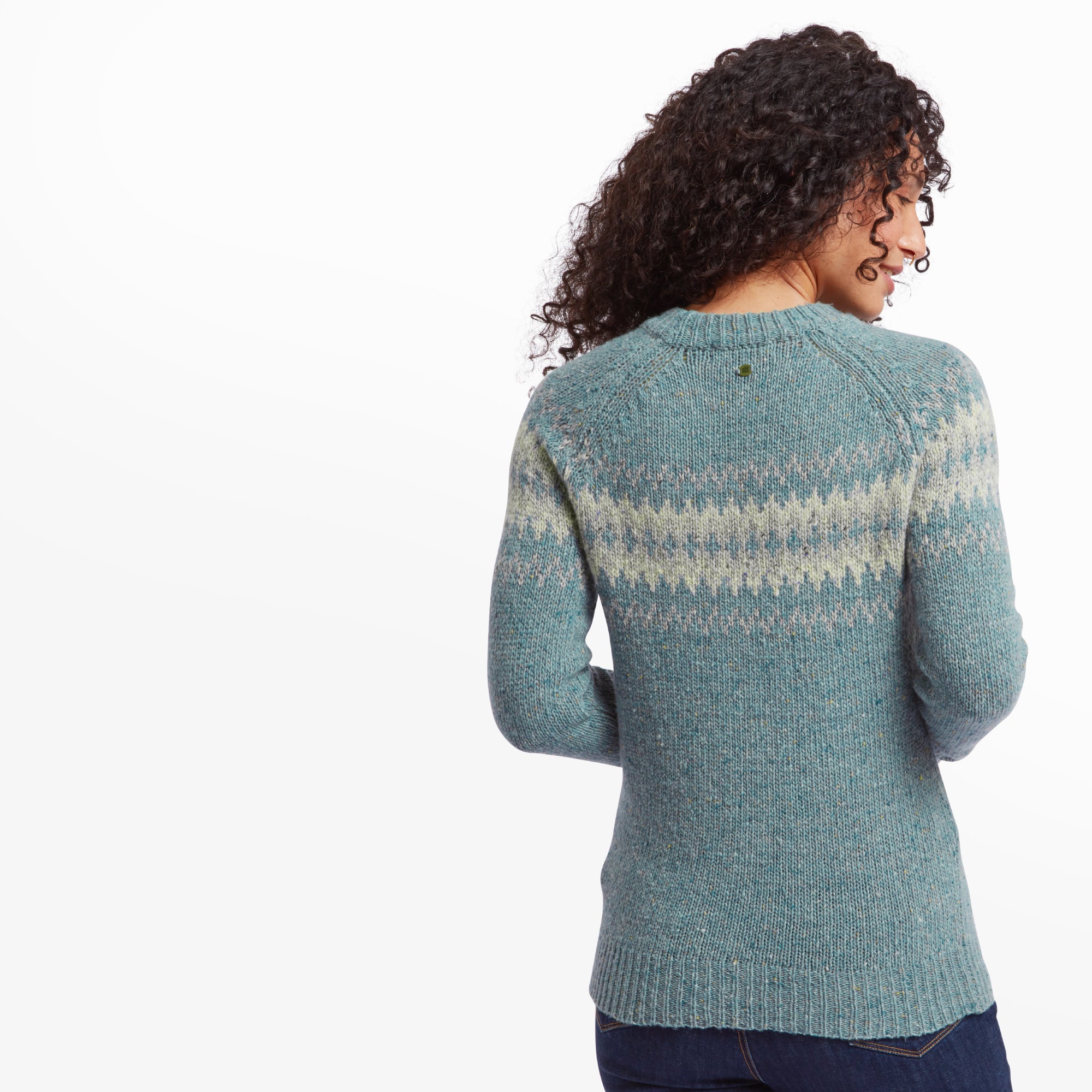 Back view of a woman wearing the Sherpa Adventure Gear Dumji Crew Sweater in Green. The fair isle pattern continues across the back of the sweater. Her natural curly hair is draped over her shoulders, and she is wearing blue jeans. The setting is a plain white background.