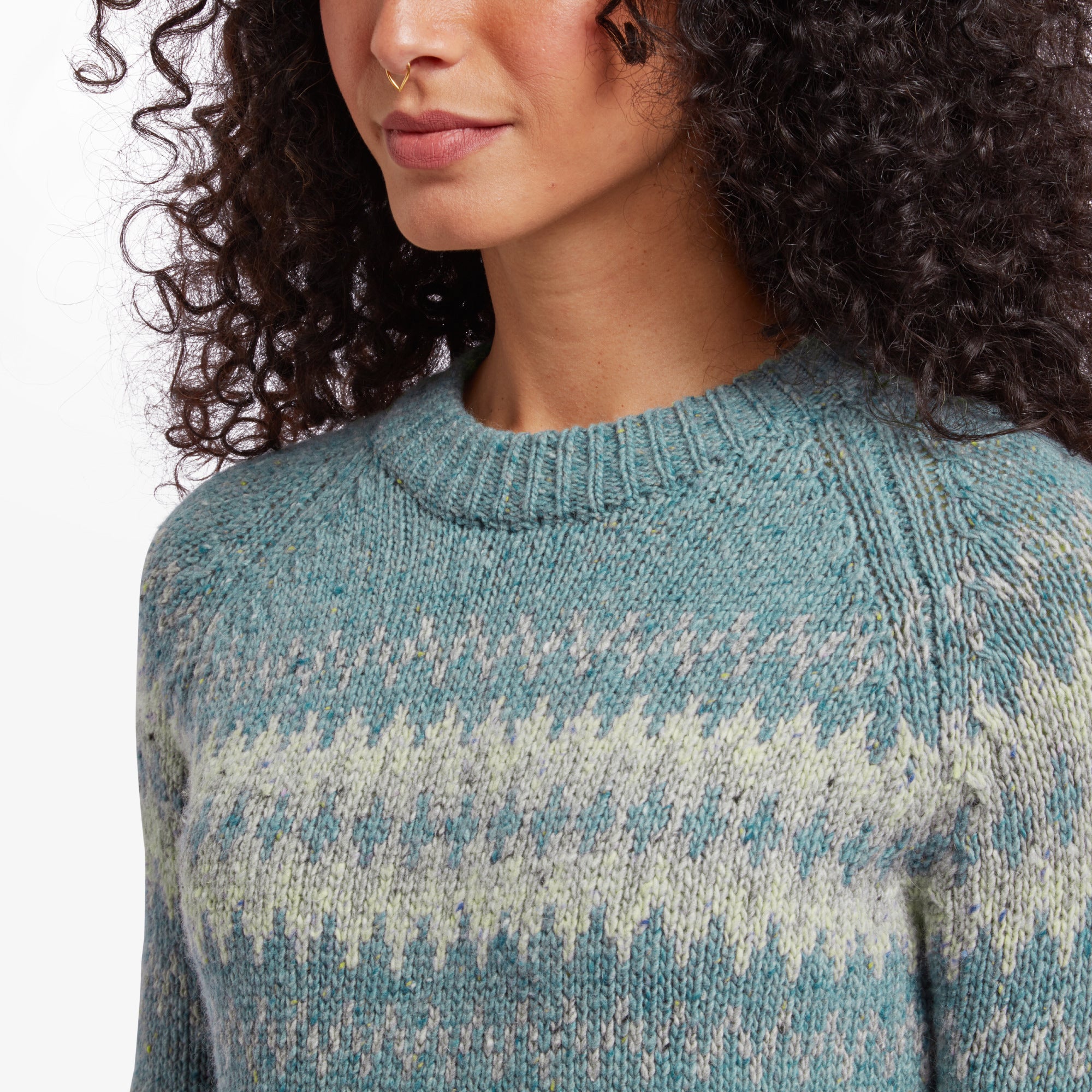 Close-up of the Sherpa Adventure Gear Dumji Crew Sweater in Green, highlighting the textured knit and fair isle design in beige and white. The crew neck and subtle detailing of the fabric are clearly visible. The woman’s face is partially shown, focusing on the sweater’s neckline and pattern.